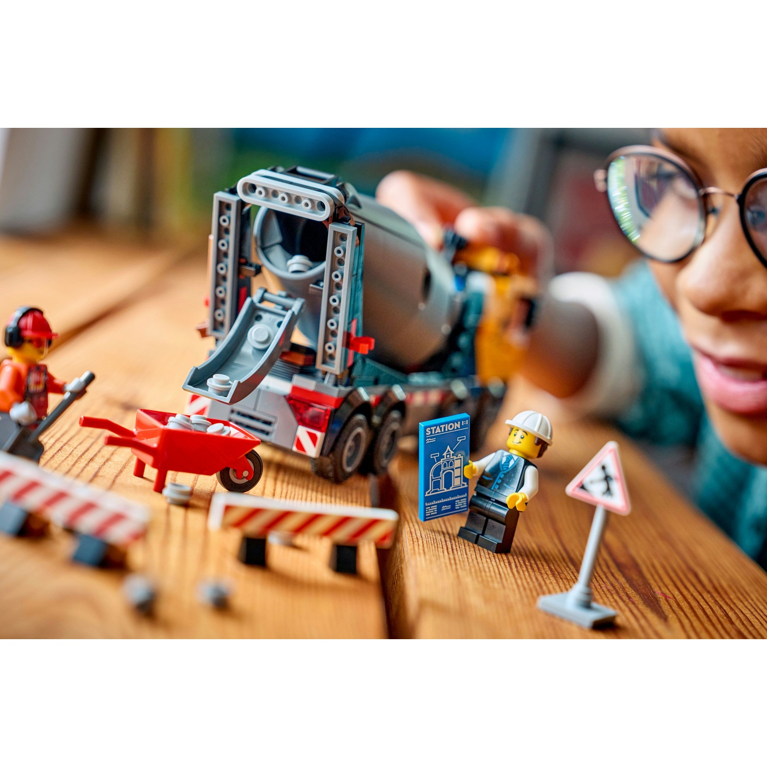 Un bambino gioca con un set di costruzioni Lego. C'è un camion betoniera, una carriola, operai edili e segnali stradali su un tavolo di legno.