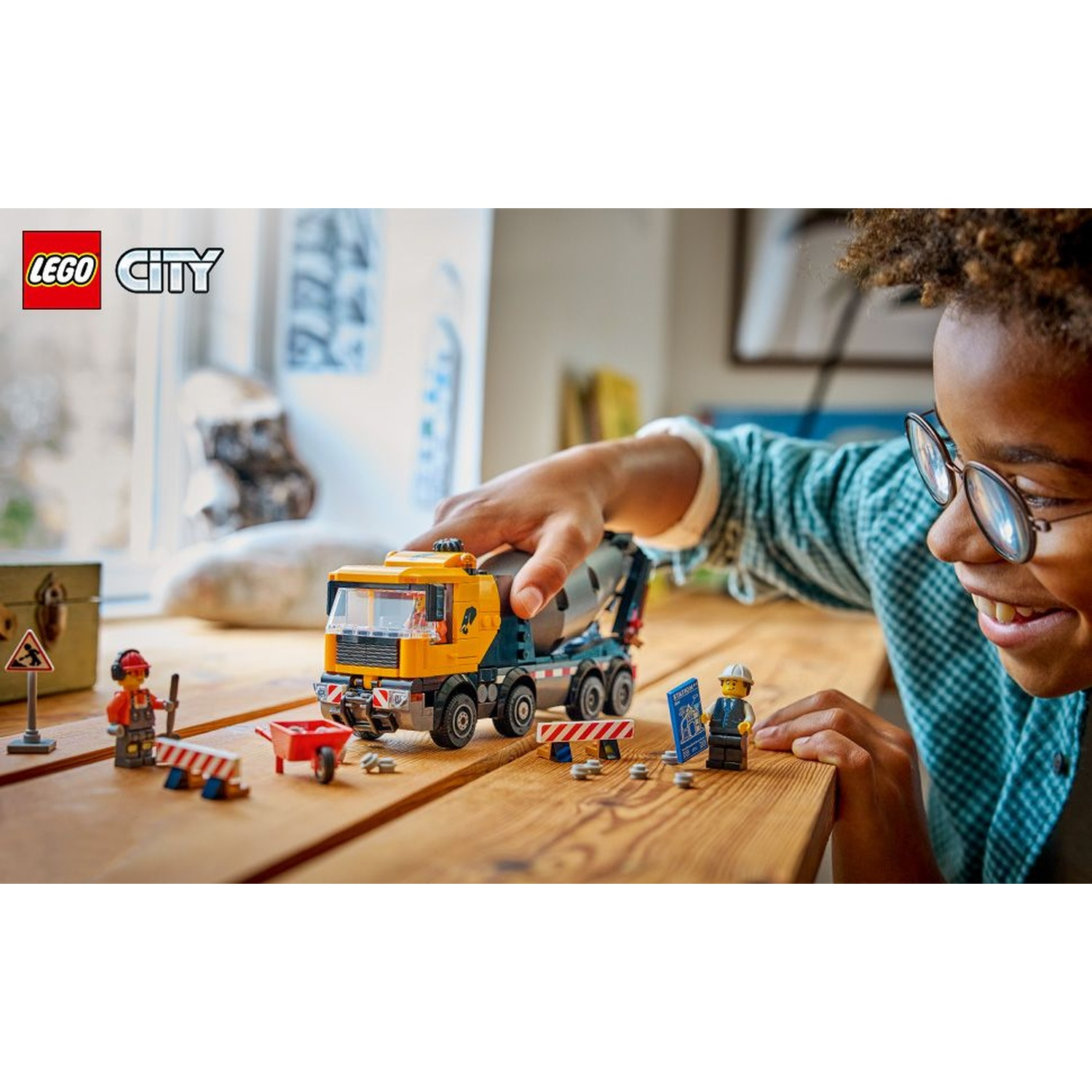 Un bambino gioca con un set di costruzioni Lego City. Un camion betoniera, figurine di operai e un segnale stradale sono su un tavolo di legno.