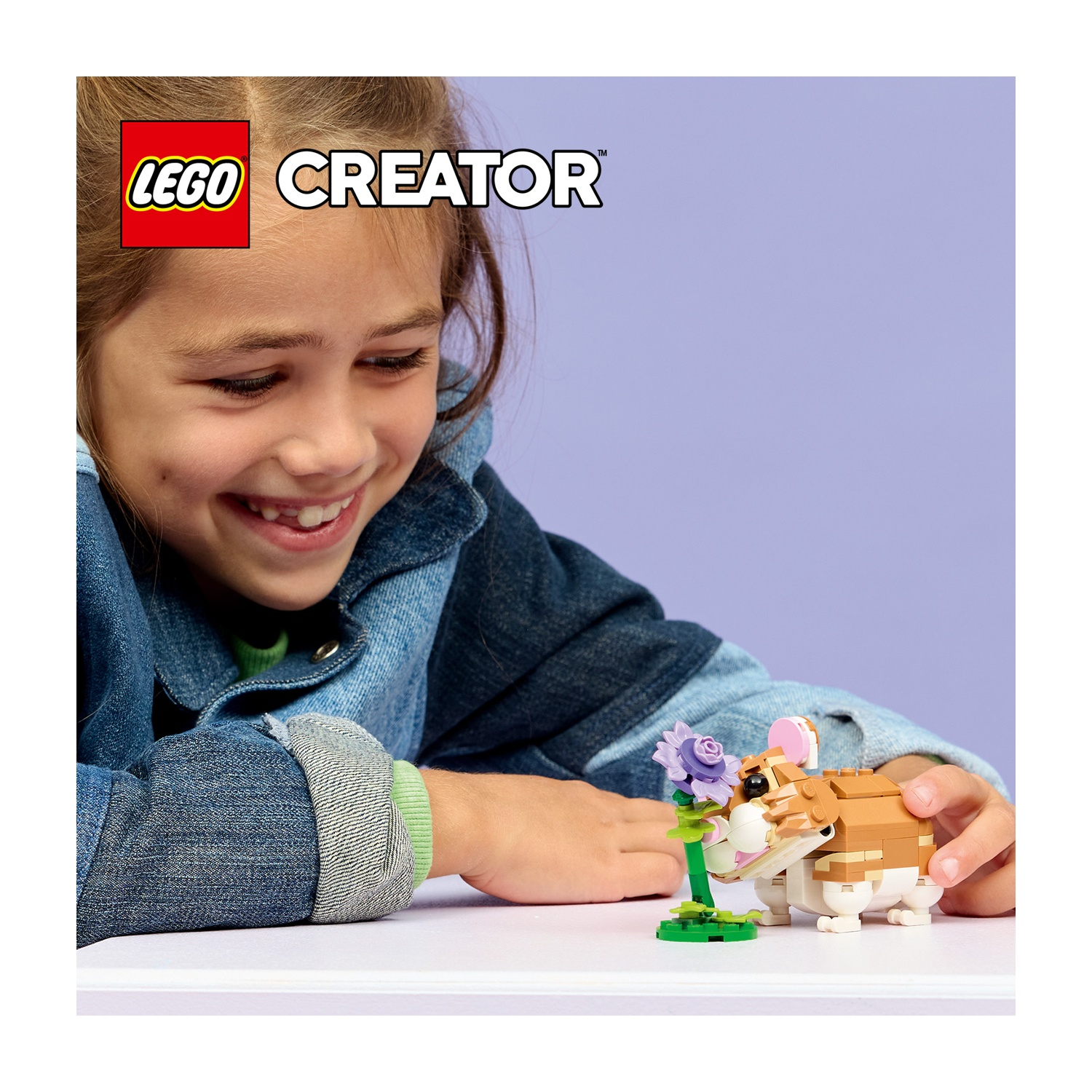 Una ragazza sorride mentre guarda un criceto e un fiore Lego. Il set Lego è su una superficie bianca, lo sfondo è viola.