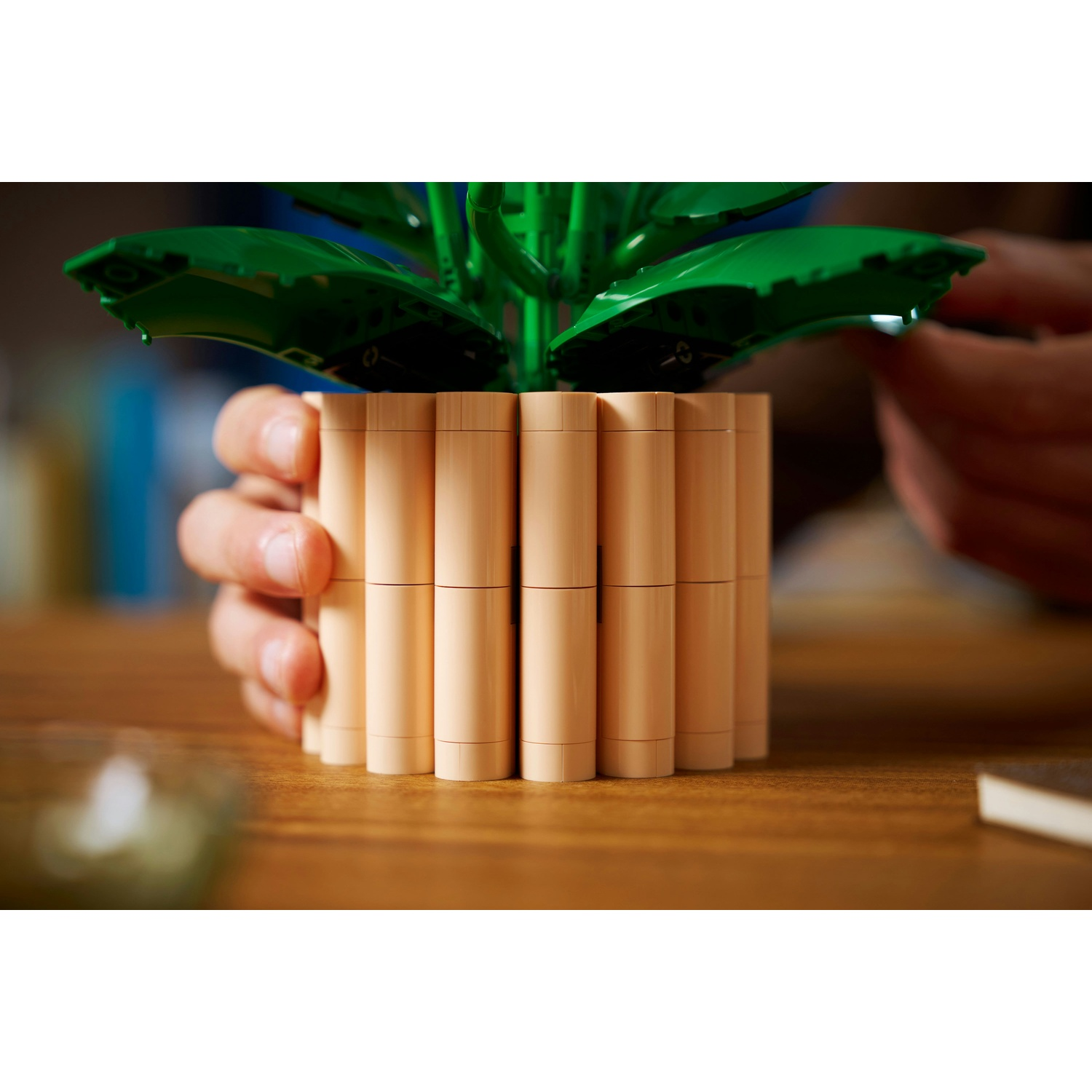 Una pianta Lego in un vaso beige è tenuta da una mano. Sopra ci sono foglie verdi. Il vaso è su un tavolo di legno.