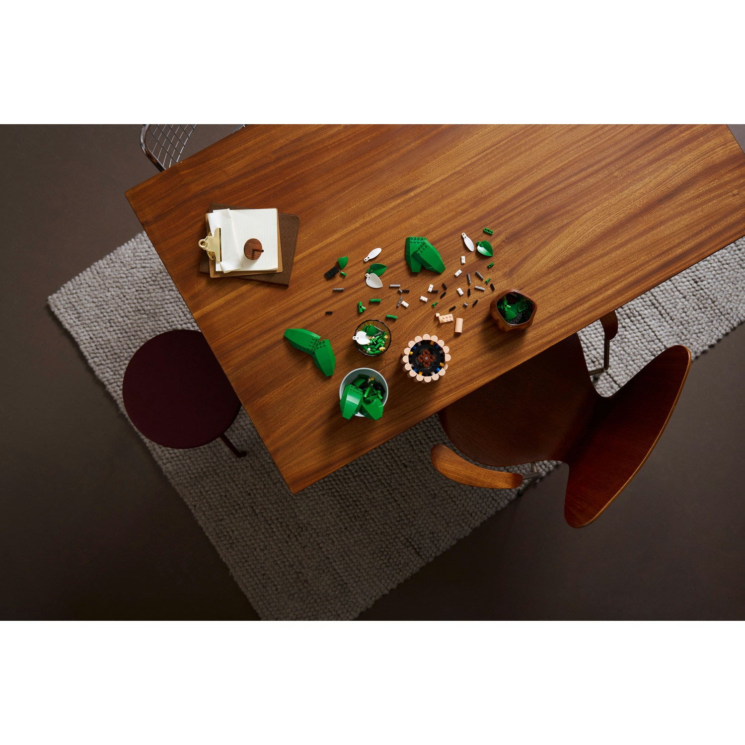 Vista dall'alto di un tavolo di legno con pezzi Lego verdi sparsi, un blocco note e una sedia rotonda. Un tappeto grigio è accanto al tavolo.