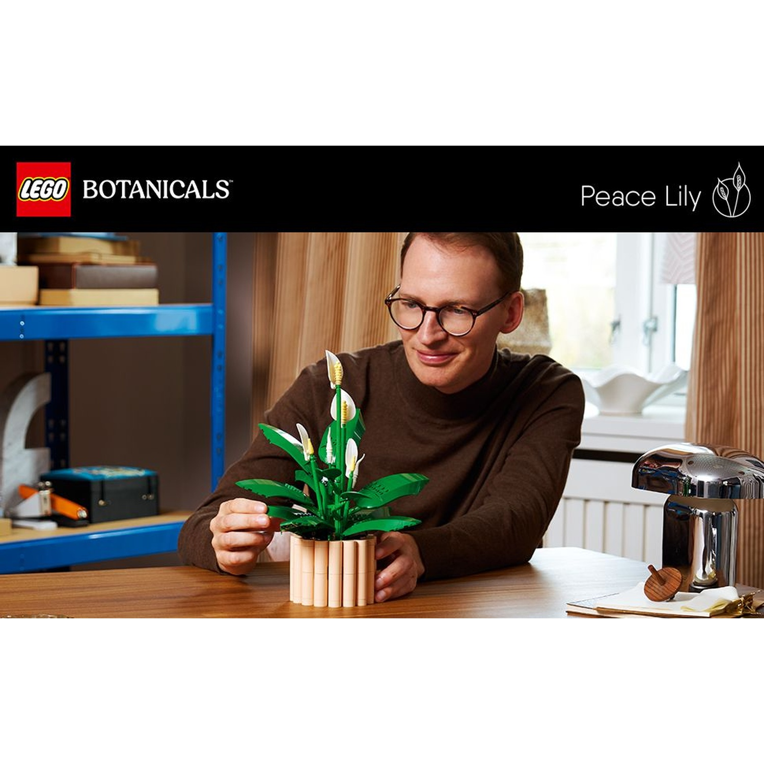 Un uomo con gli occhiali tiene una Peace Lily LEGO. È su un tavolo di legno. Una mensola e una lampada sono sullo sfondo.