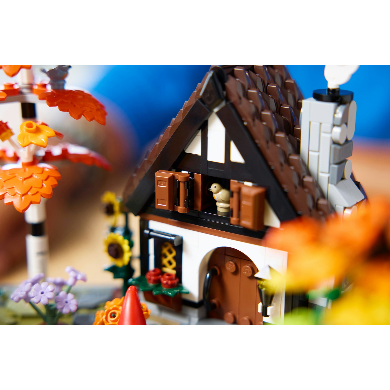 Casa Lego con tetto marrone, pareti bianche e un uccellino nella finestra. Fiori e un cono rosso in primo piano.