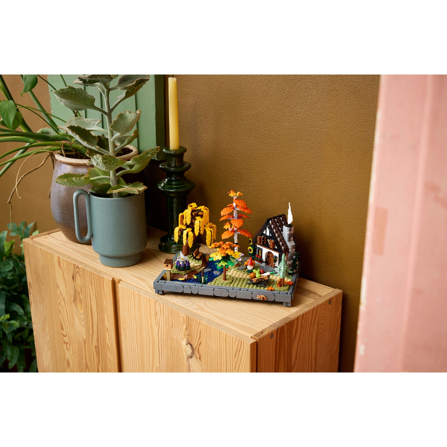 Un diorama Lego di una casa e alberi è su un mobile di legno. Piante e un candelabro sono accanto.