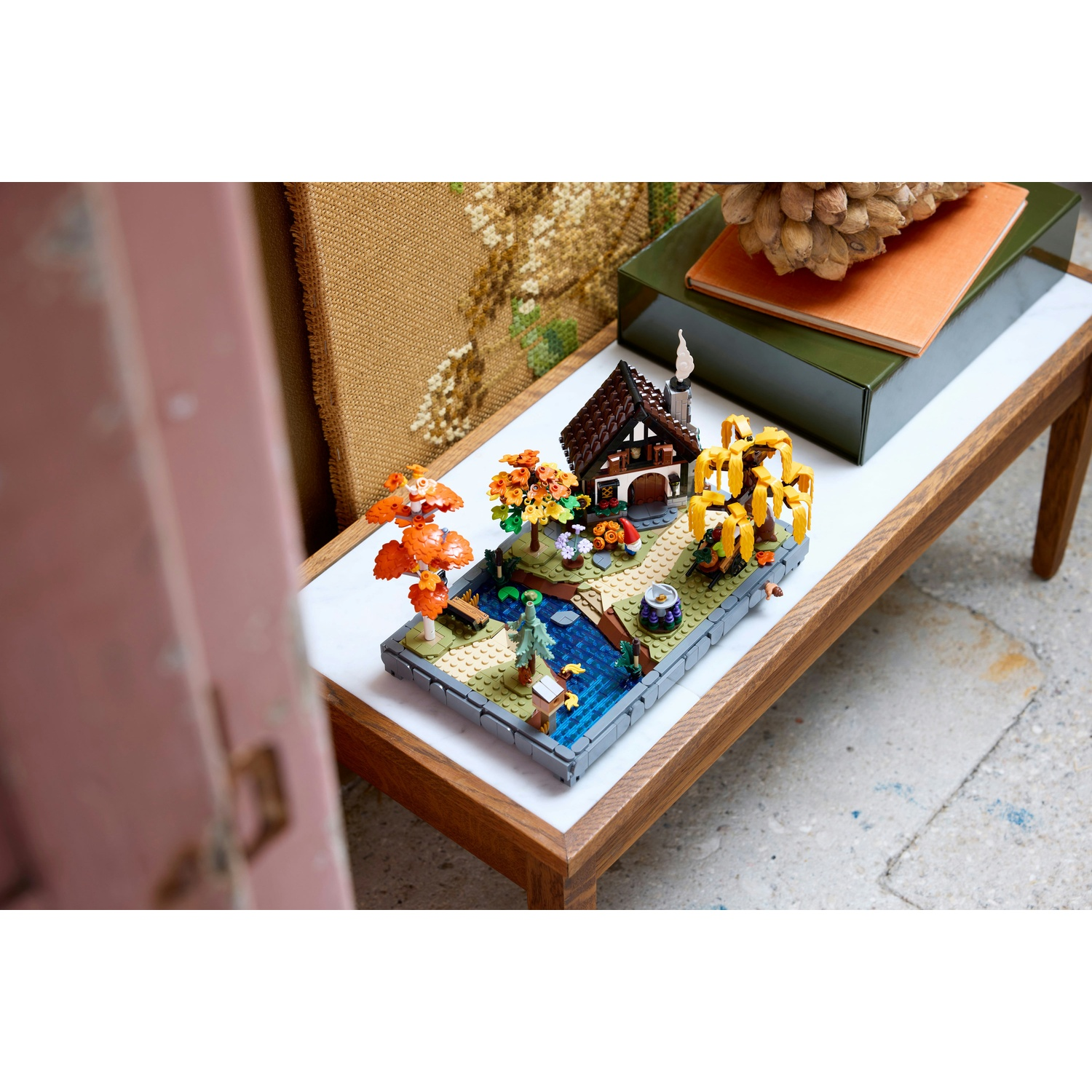 Un diorama Lego di una scena del villaggio. C'è una casa, un ruscello, alberi e altri elementi su un tavolo bianco. La scena è su un tavolo.
