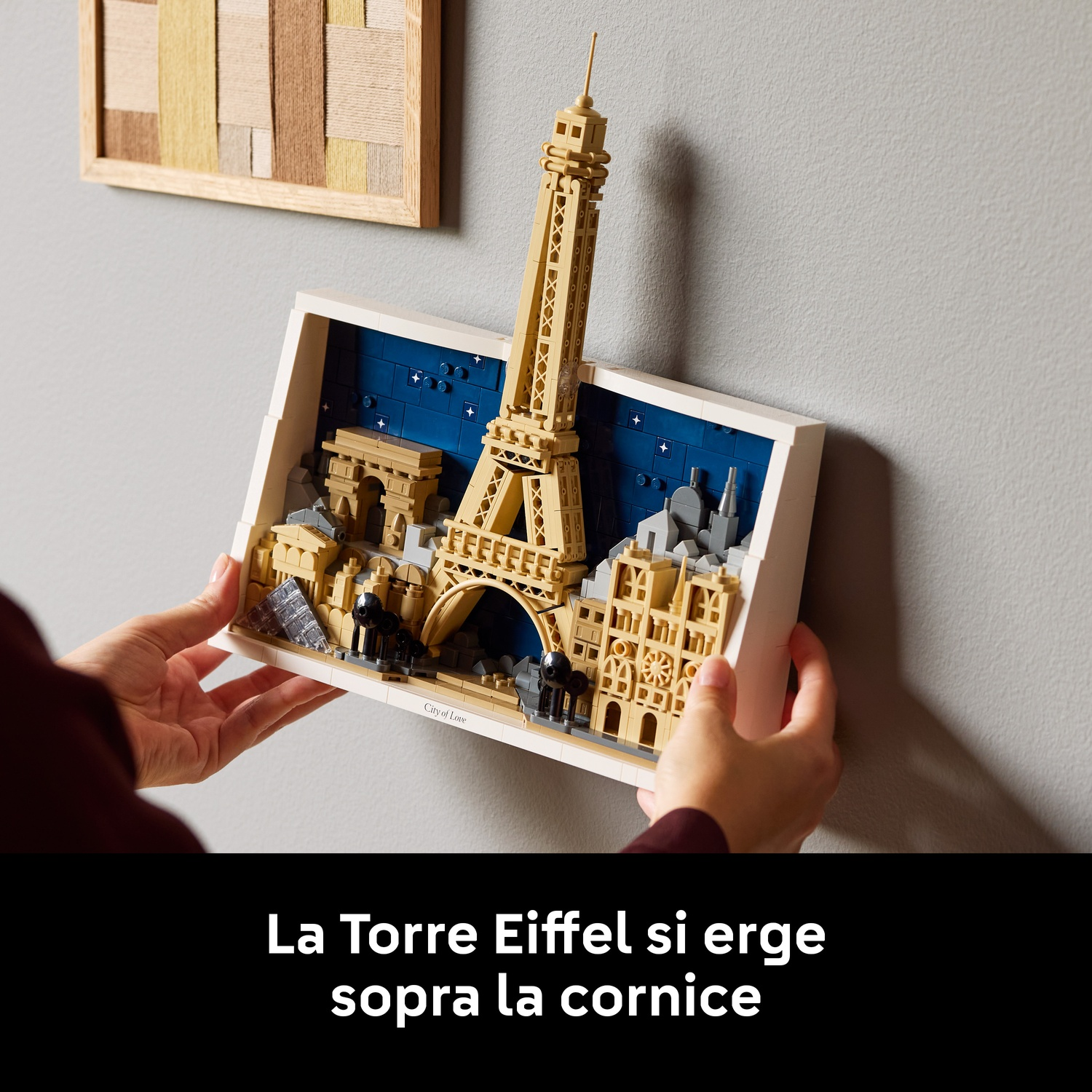 Le mani reggono un diorama Lego di Parigi con la Torre Eiffel. Un'opera d'arte incorniciata è appesa sopra. Lo sfondo è un muro grigio.