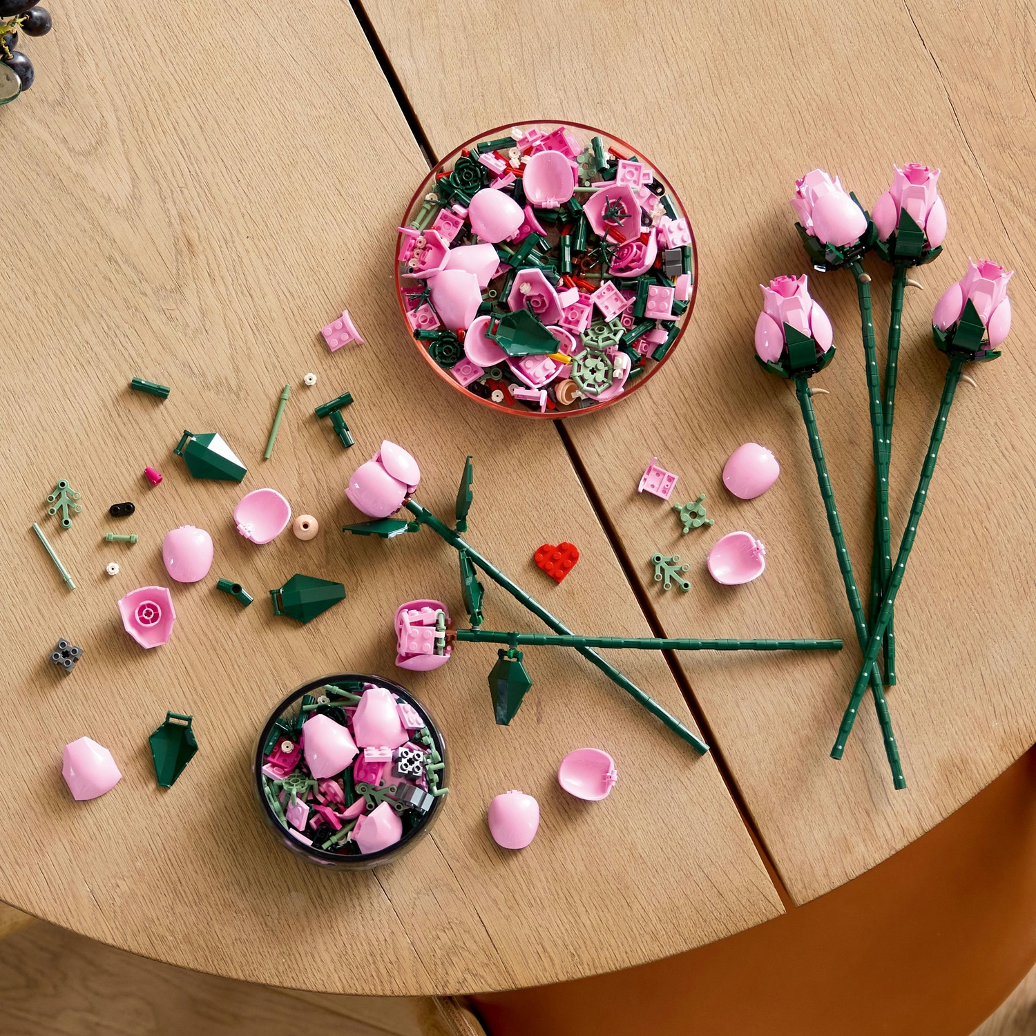 Tavolo di legno con parti di rose Lego. Petali rosa, steli verdi e rose assemblate sono sparsi intorno a due ciotole.