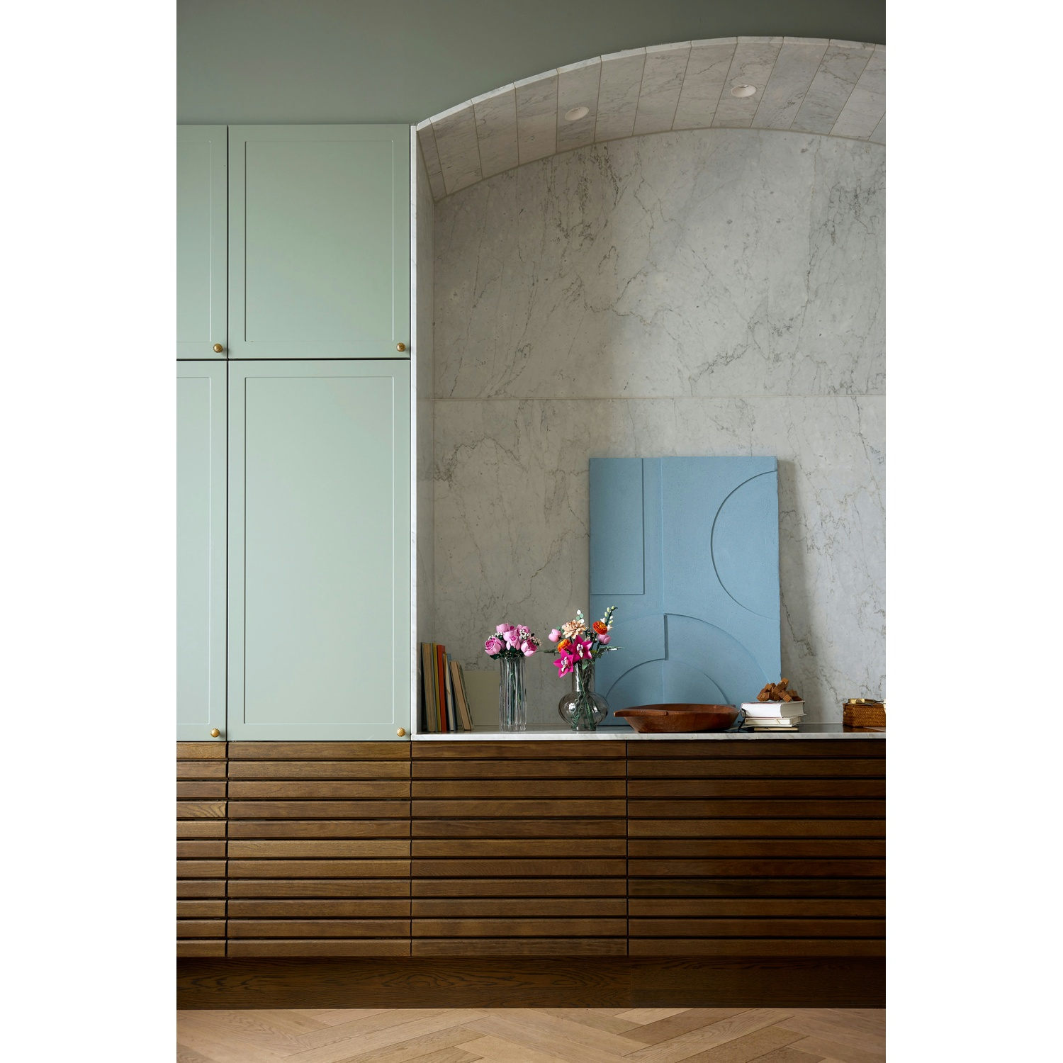 Un interno moderno con armadi verdi, cassetti in legno, parete in marmo e un'opera d'arte blu. Sono presenti anche fiori e libri.