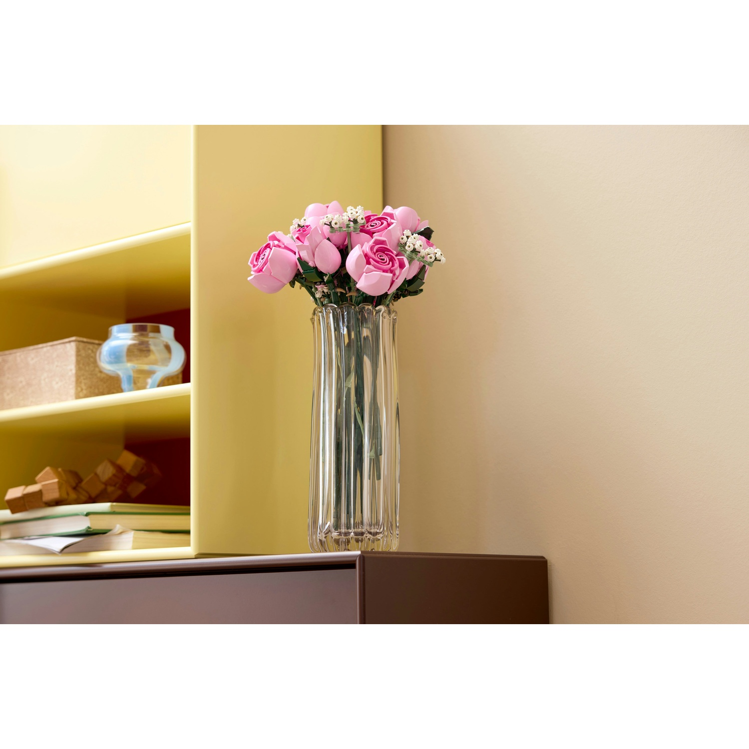 Un vaso di vetro trasparente con rose rosa poggia su una superficie marrone scuro. Mensole gialle contengono oggetti. Parete beige sullo sfondo.