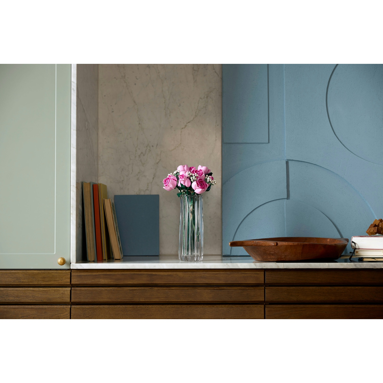 Un vaso di fiori rosa su una superficie di marmo bianco. Libri, una ciotola di legno e una parete blu con motivo sono visibili.