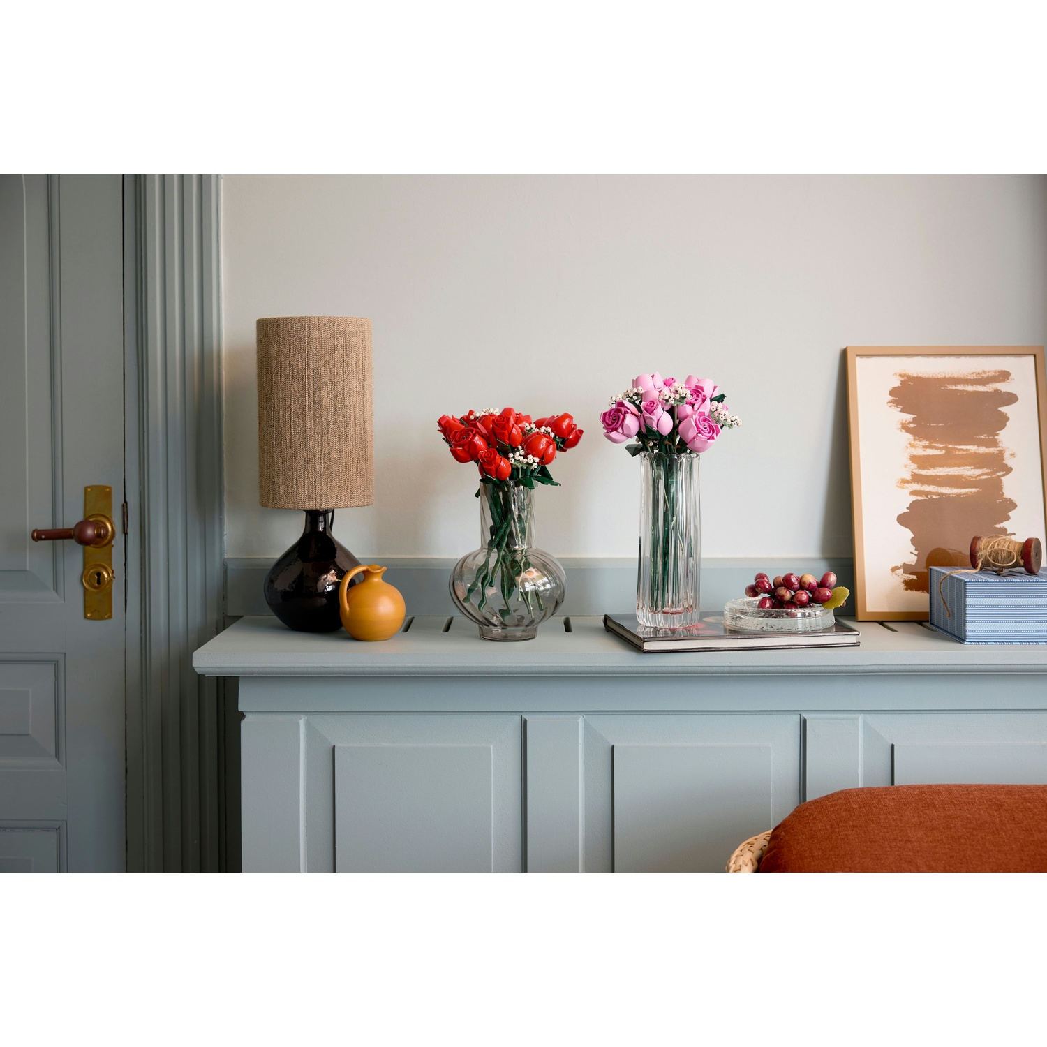 Una stanza con un mobile blu che contiene vasi di fiori rossi e rosa. Una lampada, un quadro e altri oggetti sono sul mobile.