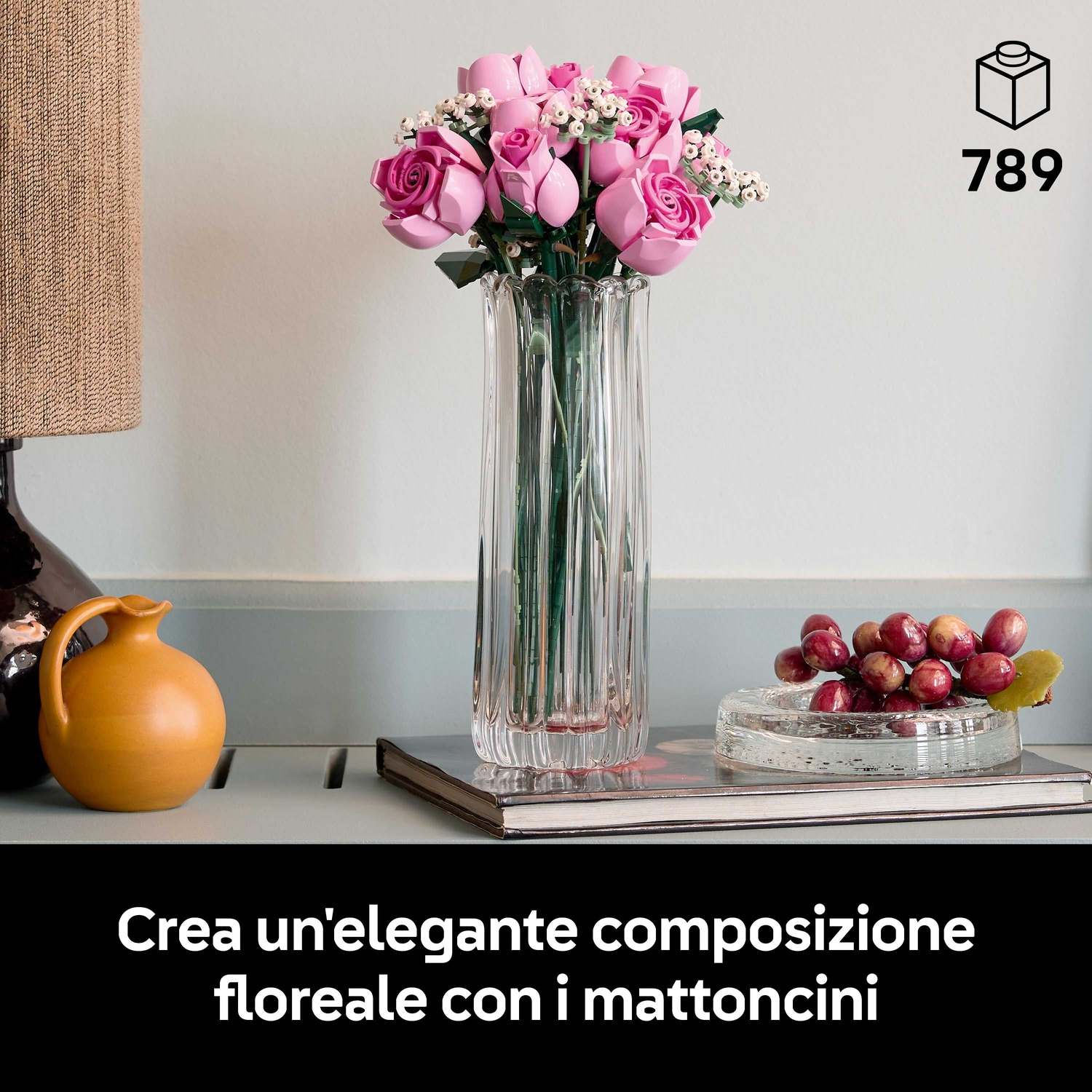 Un vaso trasparente con rose Lego rosa. Una brocca gialla e uva in un piatto di vetro. Una lampada a sinistra.