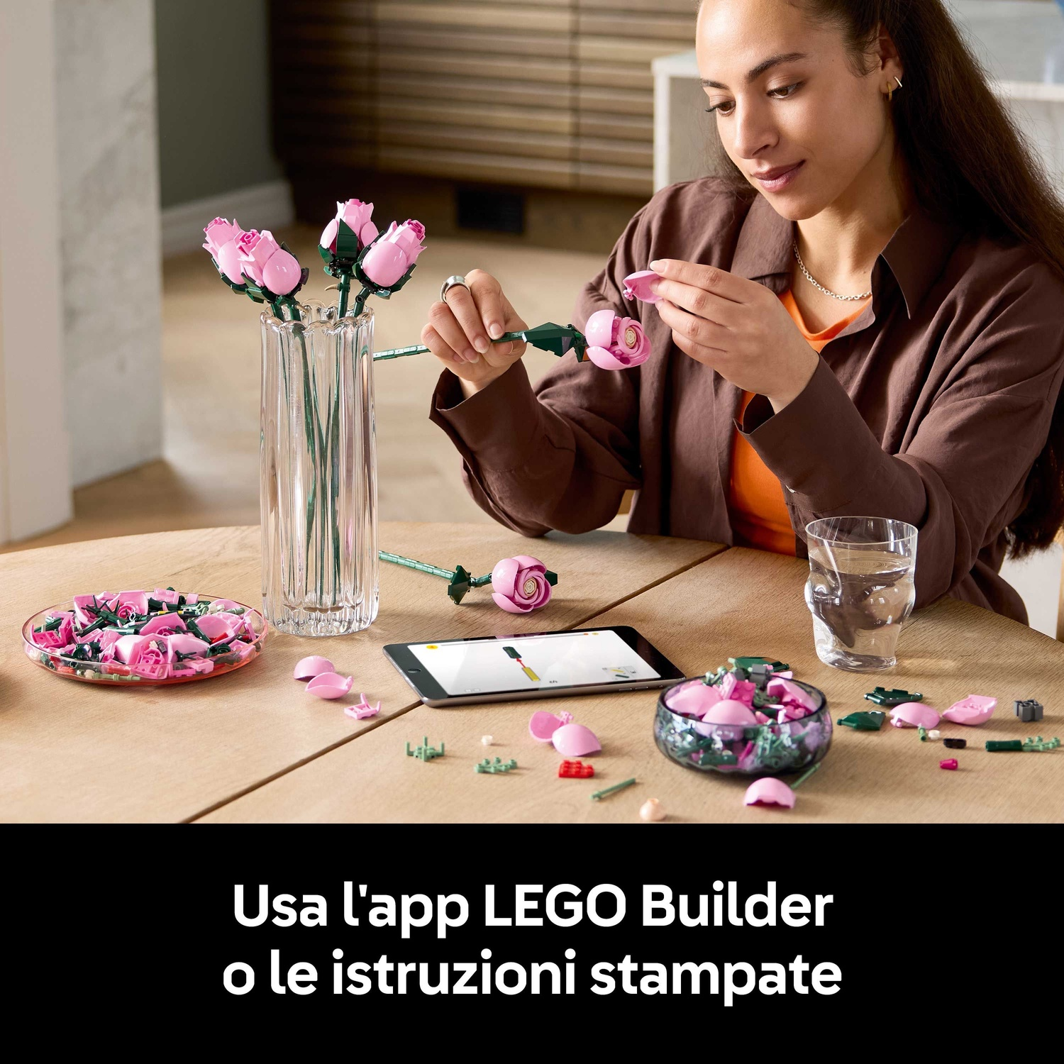 Donna che assembla rose LEGO. Un vaso con rose, un tablet e un bicchiere d'acqua sono sul tavolo. Parti sparse.