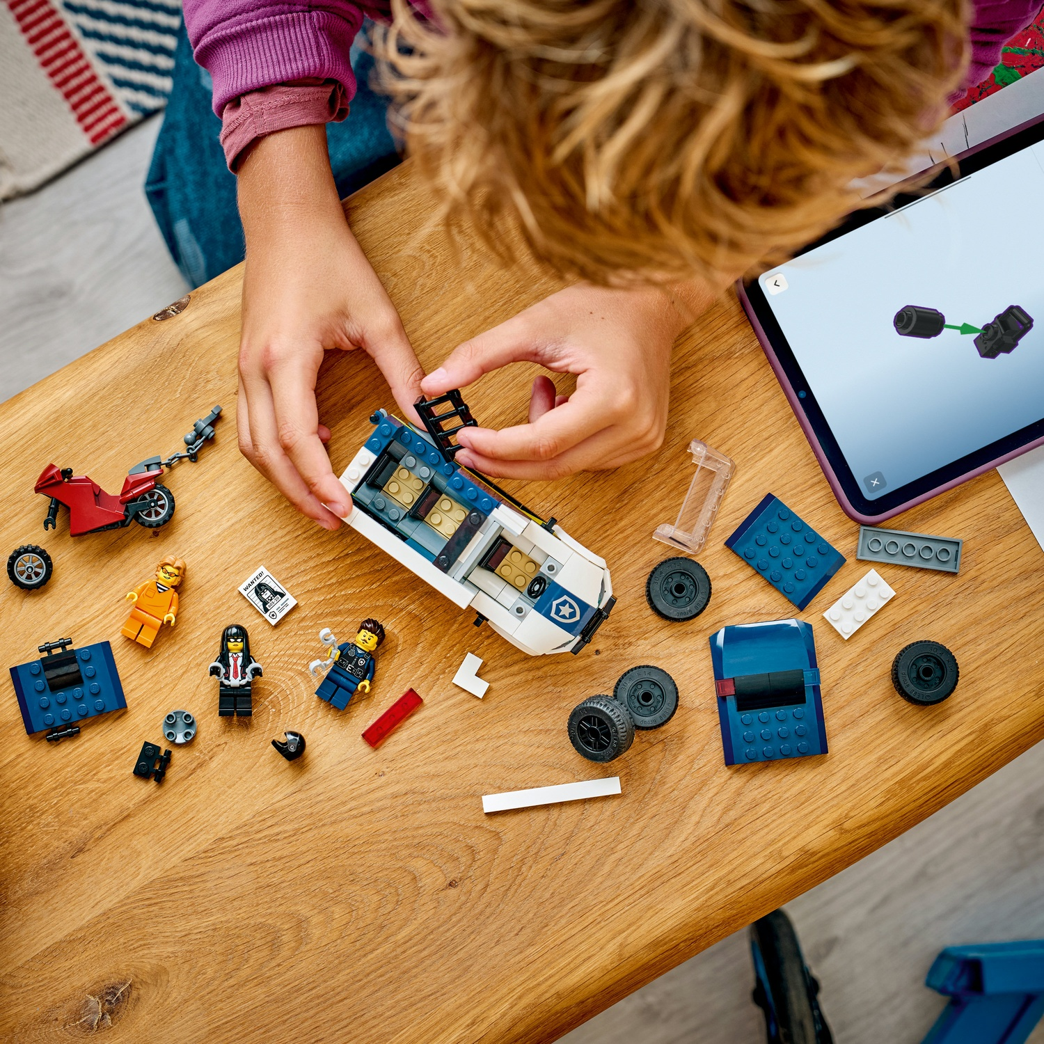 Un bambino assembla un'auto della polizia Lego su un tavolo di legno. Figure e pezzi Lego sono sparsi, insieme a un tablet.