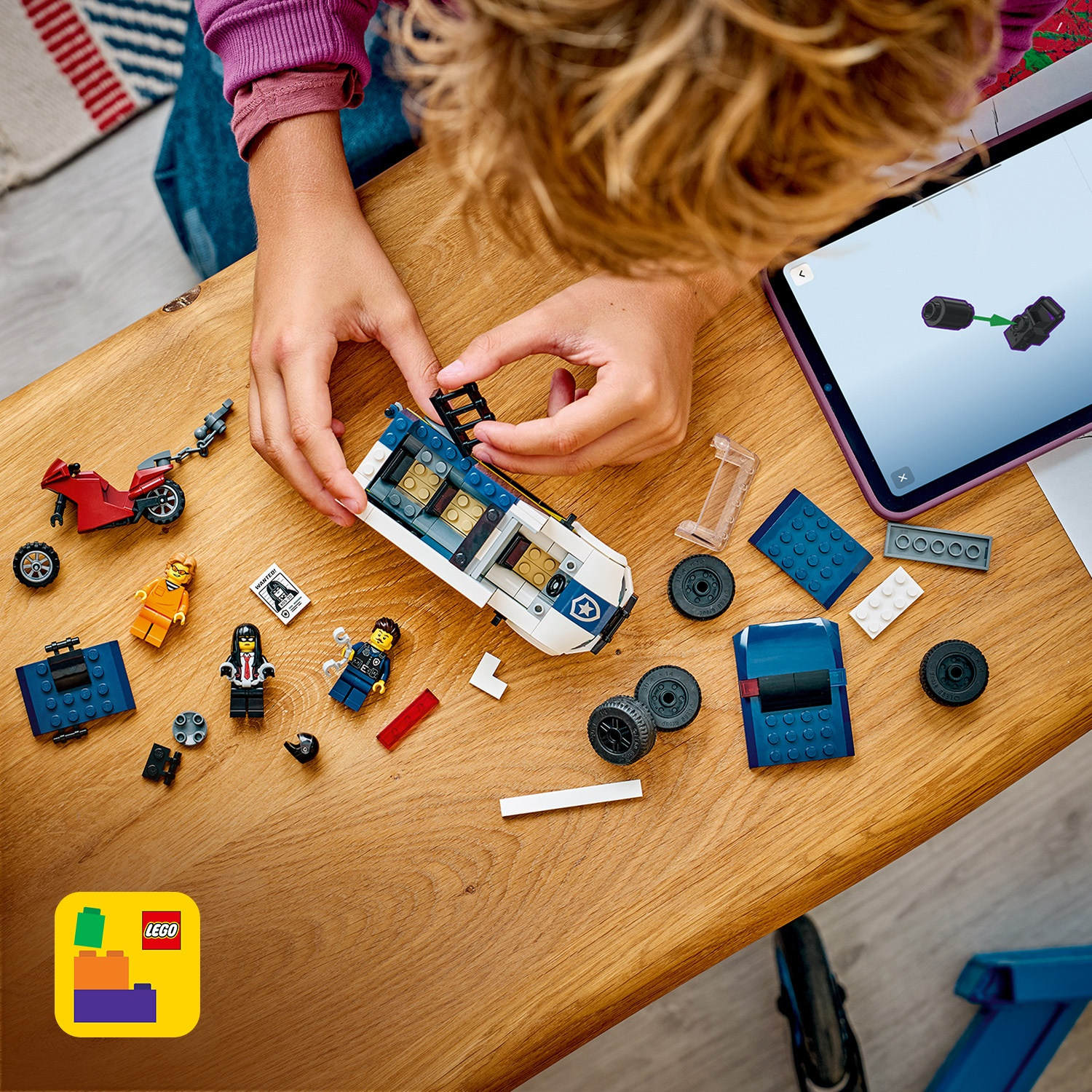 Un bambino assembla un'auto della polizia Lego su un tavolo di legno. Sono visibili pezzi Lego, personaggi, una moto rossa e un tablet.