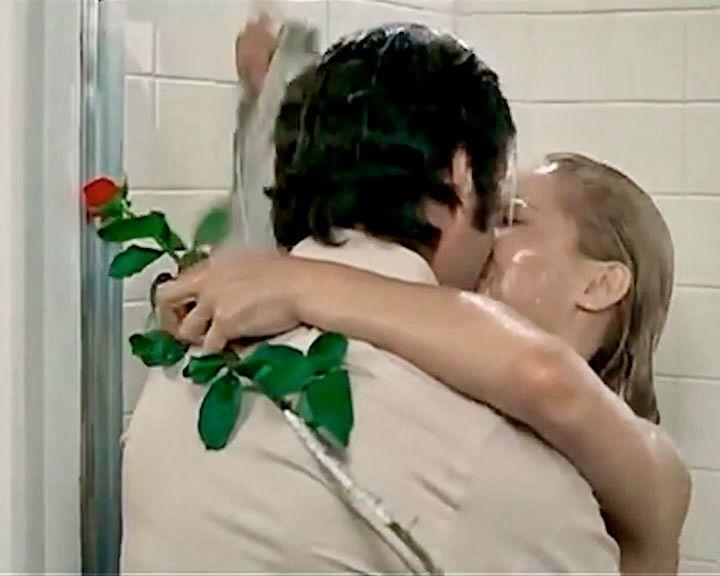 Ein Paar küsst sich unter der Dusche. Ein Mann hält eine Rose. Die Frau hat nasses blondes Haar. Weiße Fliesen im Hintergrund.