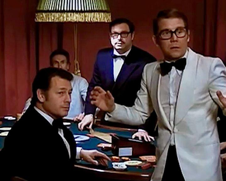 Männer in Smokings an einem Pokertisch. Ein Mann im weißen Jackett gestikuliert. Andere schauen zu, einige tragen Brillen. Chips und Karten sichtbar.