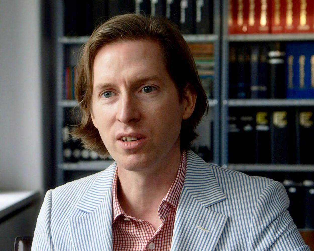 Ein Mann mit hellem Haar und blauen Augen, der einen gestreiften Blazer über einem karierten Hemd trägt, steht vor einem Bücherregal.