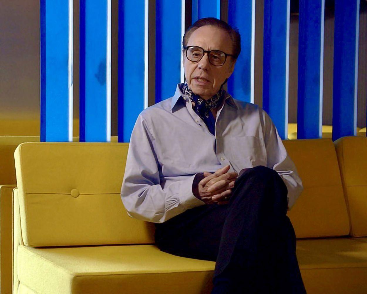 Ein älterer Mann mit Brille und Schal sitzt auf einem gelben Sofa. Blau-weiße Paneele sind im Hintergrund.