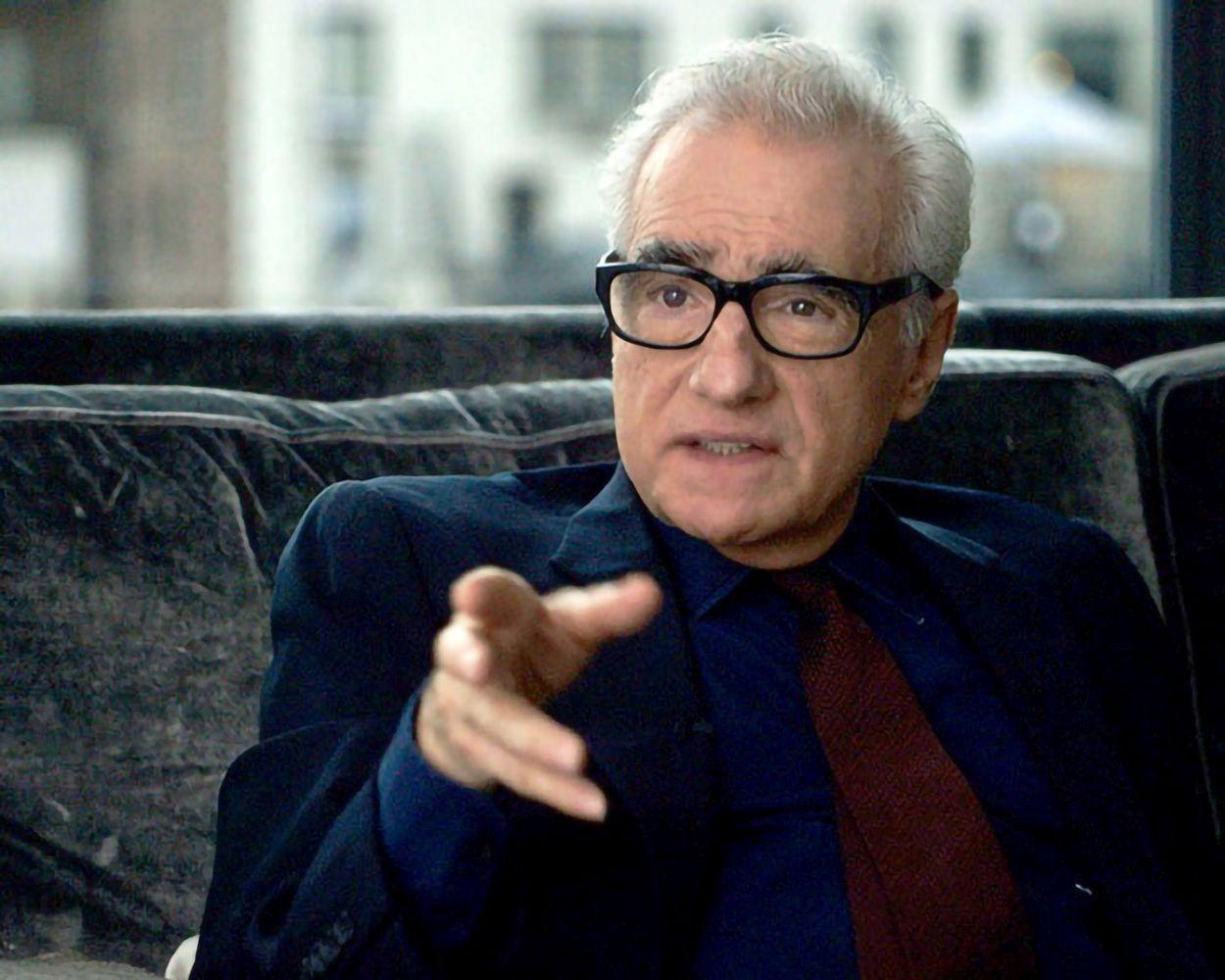 Ein älterer Mann mit Brille zeigt. Er trägt einen dunklen Anzug, ein blaues Hemd und eine rote Krawatte. Er sitzt auf einem dunklen Sofa.