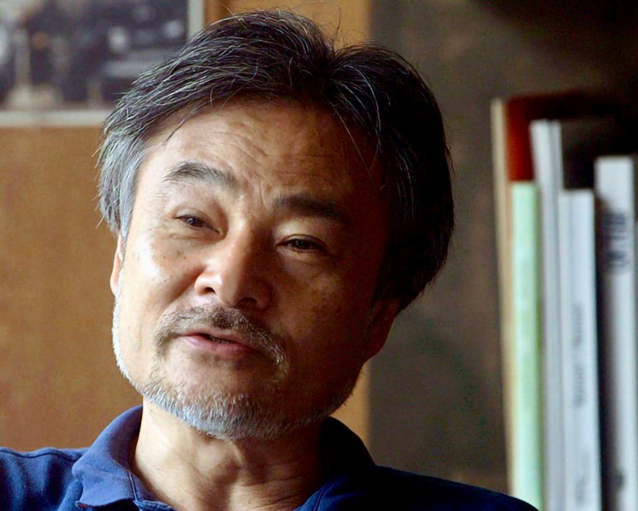 Ein älterer asiatischer Mann mit grau werdendem Haar und Schnurrbart lächelt. Er trägt ein blaues Hemd. Bücherregal im Hintergrund.