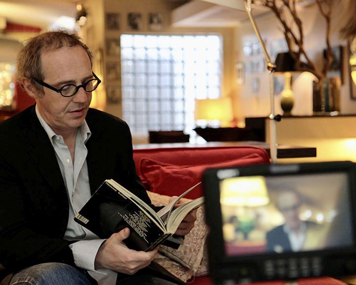 Ein Mann mit Brille liest ein Buch auf einem roten Sofa. Ein Bildschirm zeigt sein verschwommenes Bild. Hintergrund: Fenster, Lampe.