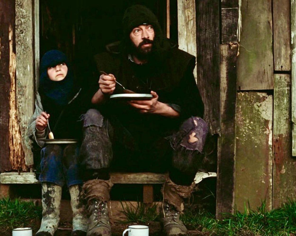 Zwei Personen essen vor einer Holzkonstruktion. Sie tragen zerrissene Kleidung und Stiefel. Zwei weiße Tassen stehen auf dem Boden, Gras davor.
