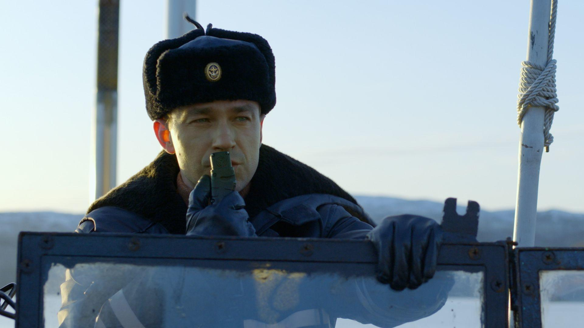 Ein Mann in Uniform und Mütze hält ein Gerät und schaut hinaus. Schneeiger Hintergrund und Metallstangen umrahmen die Sicht. Klarer Himmel.
