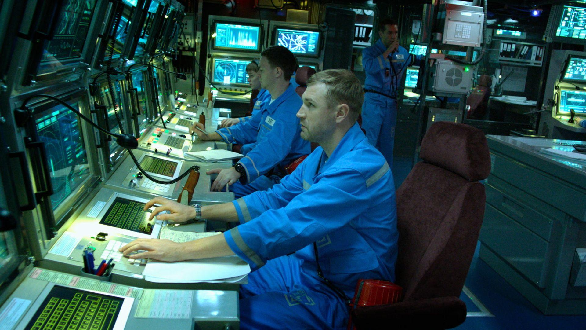 Männer in blauen Uniformen bedienen Computer in einem Kontrollraum. Mehrere Bildschirme zeigen Daten. Ein Mann spricht über ein Headset.