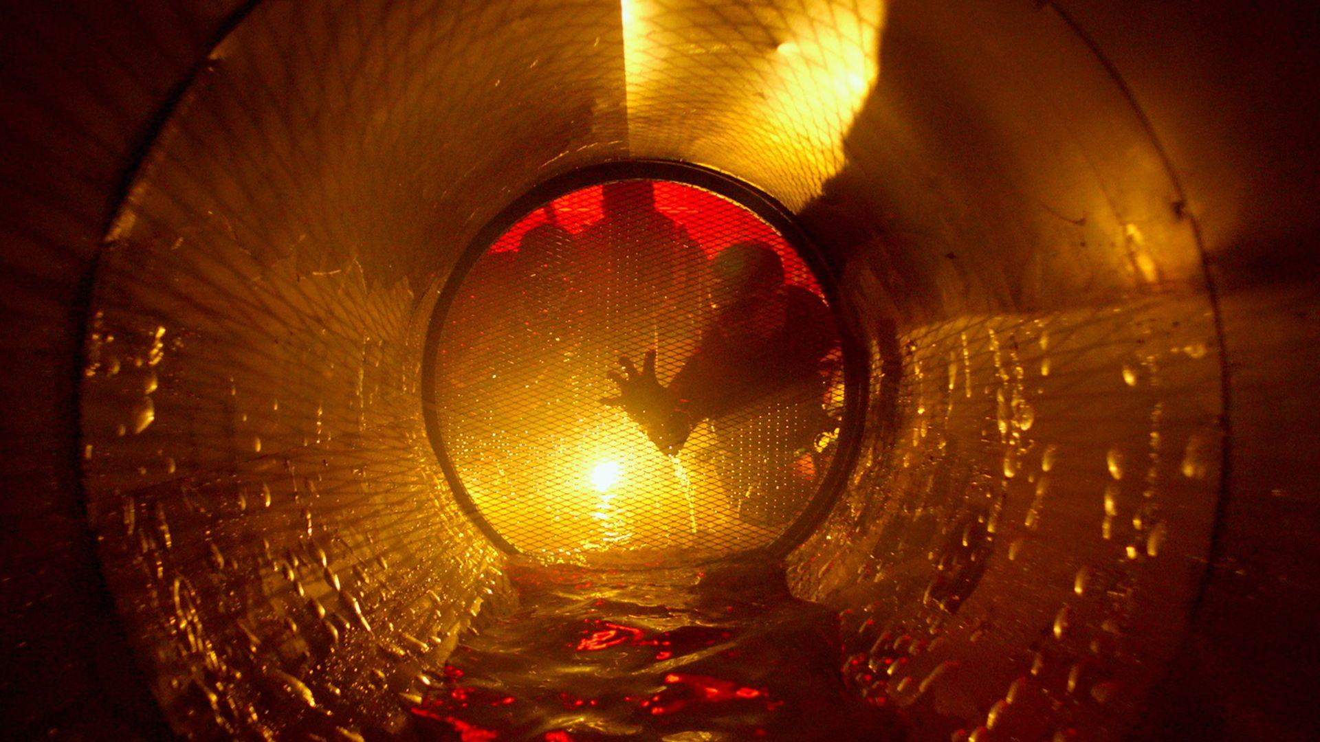 In einem kreisförmigen Tunnel greift eine Hand durch ein Metallgitter. Die Szene wird von einem hellen, warmen Licht beleuchtet.