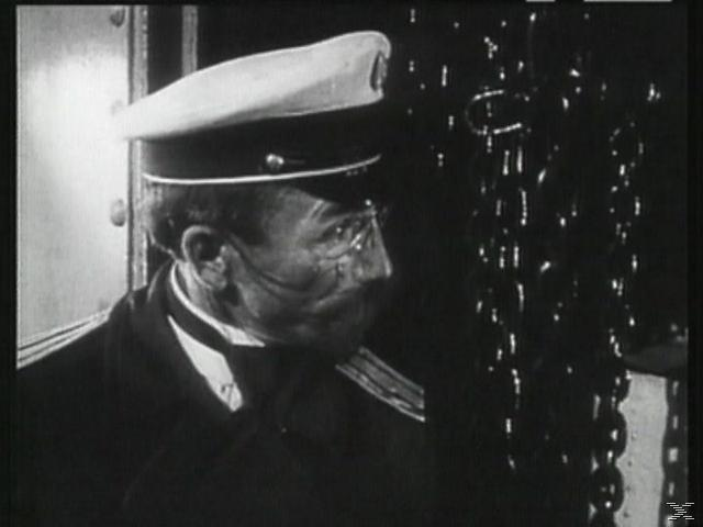 Ein Mann mit weißer Mütze und Uniform blickt auf eine Kette. Das Bild ist schwarzweiß, aus einem Film.
