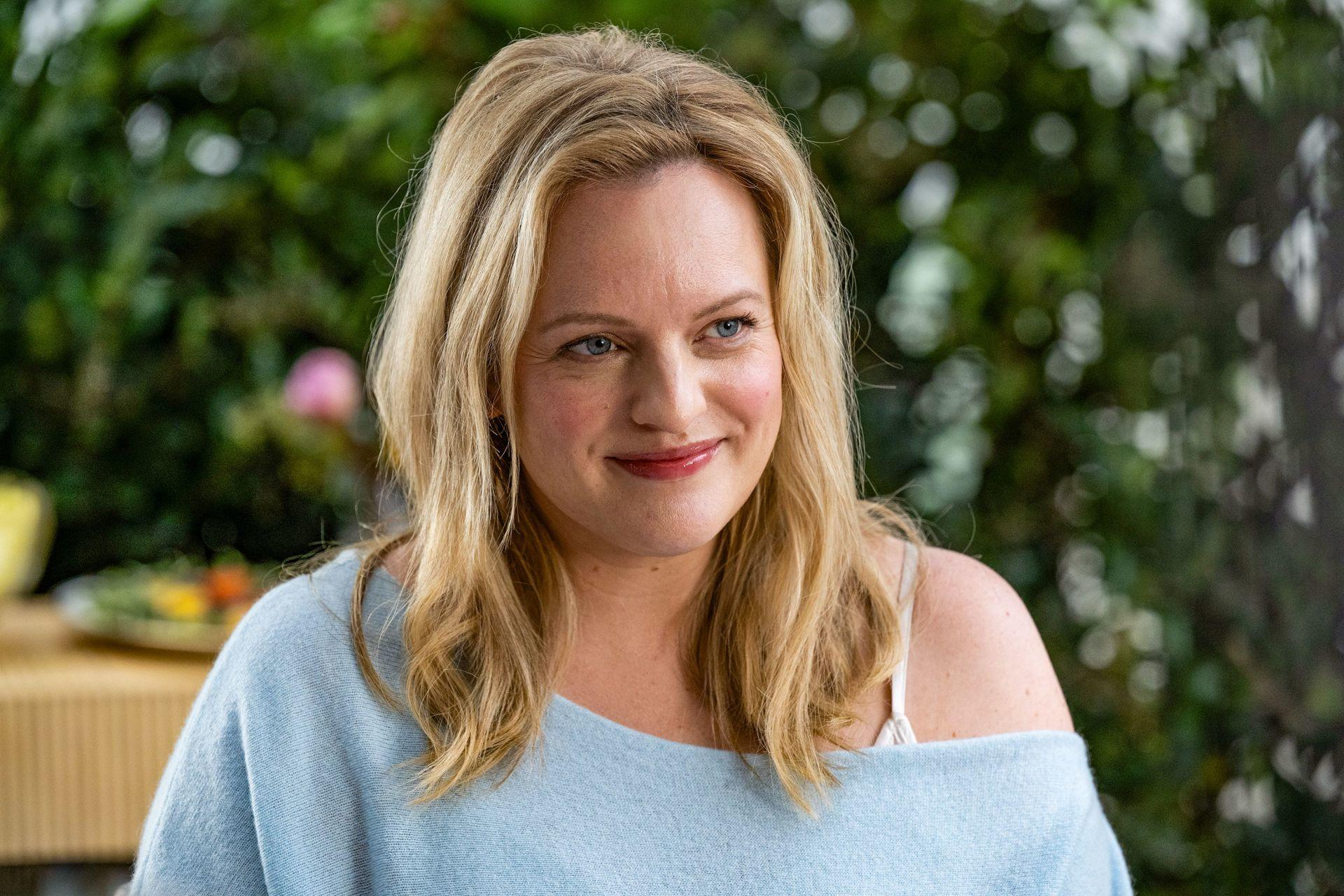 Eine Frau mit blonden Haaren, blauen Augen und einem hellblauen Pullover lächelt. Der Hintergrund ist verschwommenes Grün.