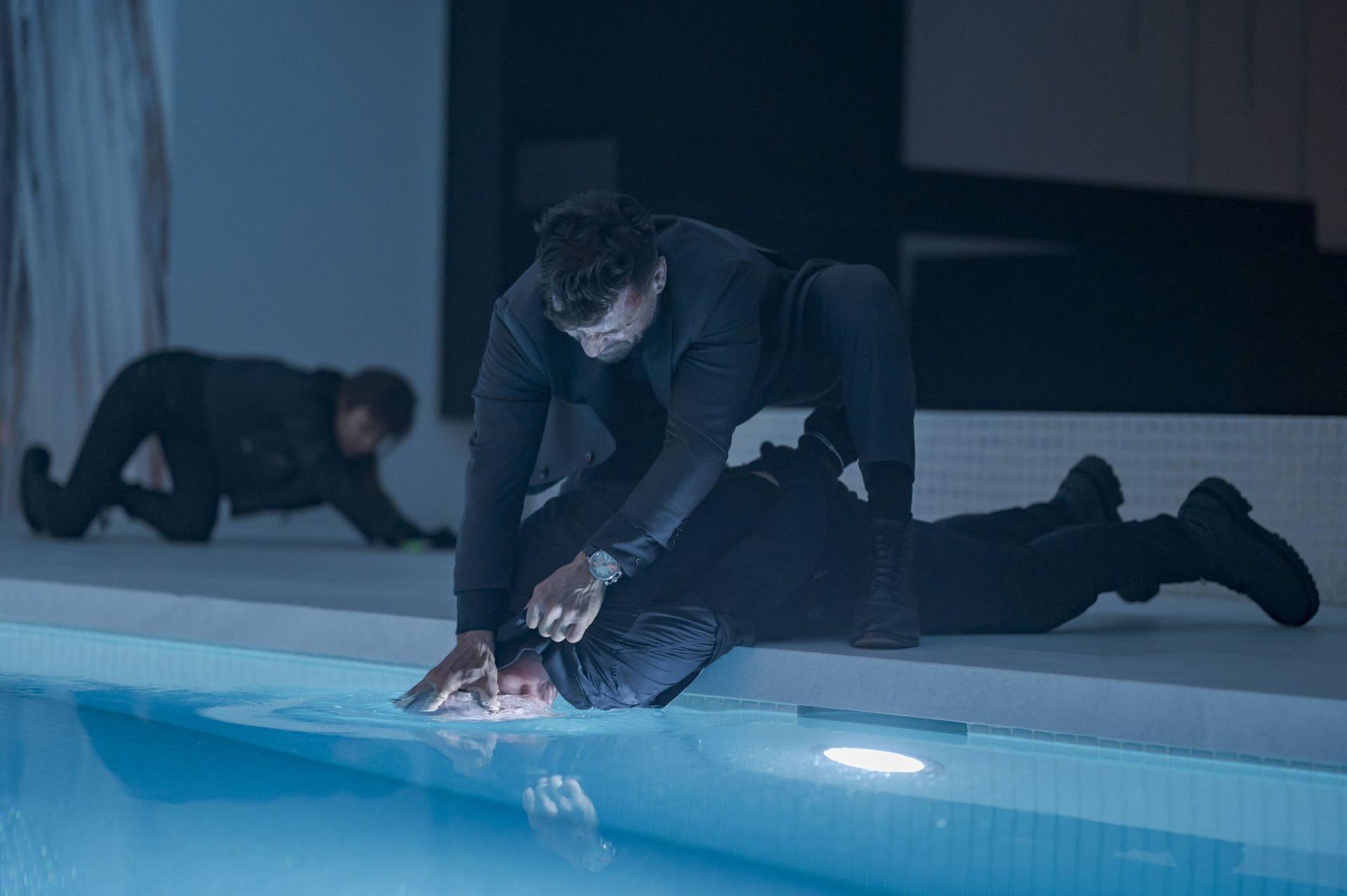 Zwei Männer in einem Pool. Einer ist im Wasser untergetaucht, der andere hält ihn fest. Eine dritte Figur kriecht auf dem Deck.