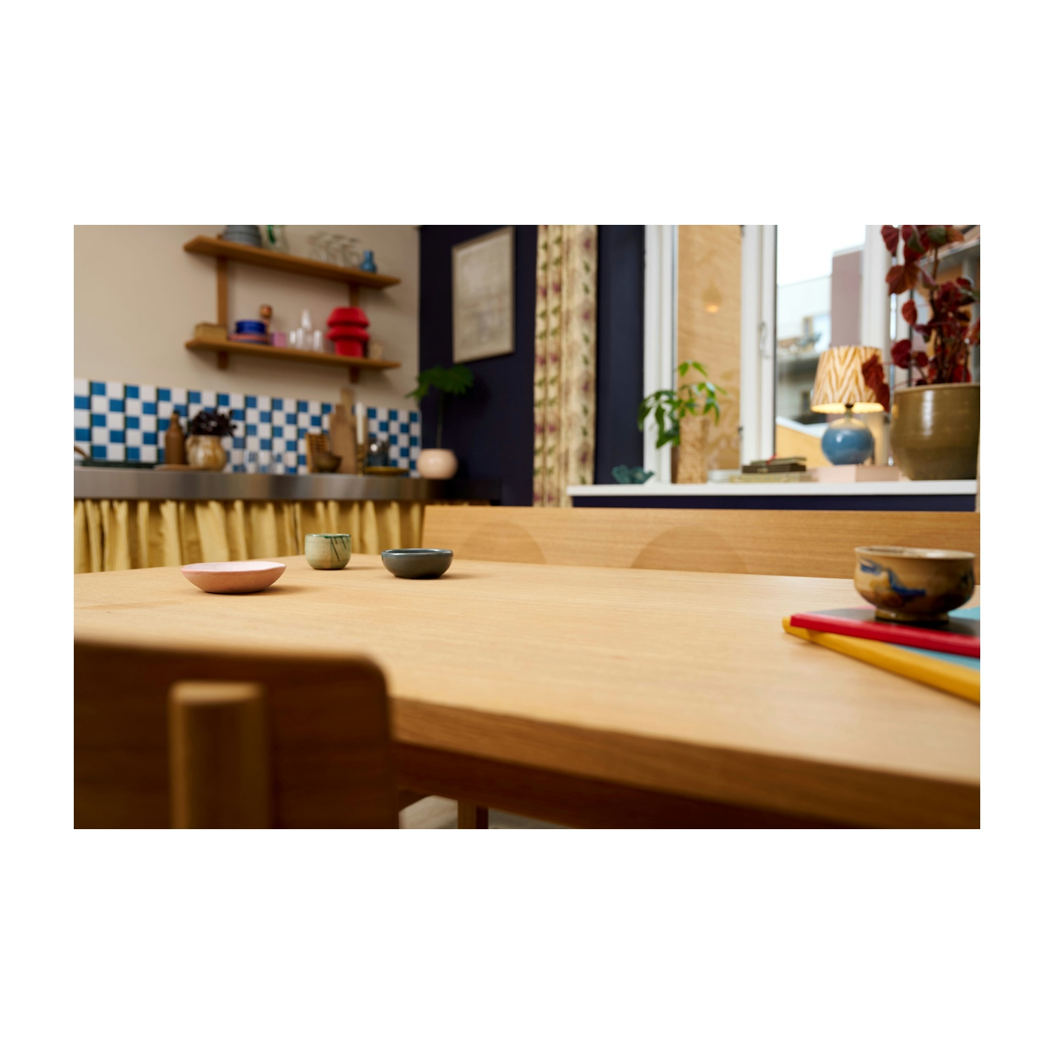 Tavolo in legno con tre piccole ciotole e libri. Lo sfondo comprende cucina, finestra, piante e una lampada.