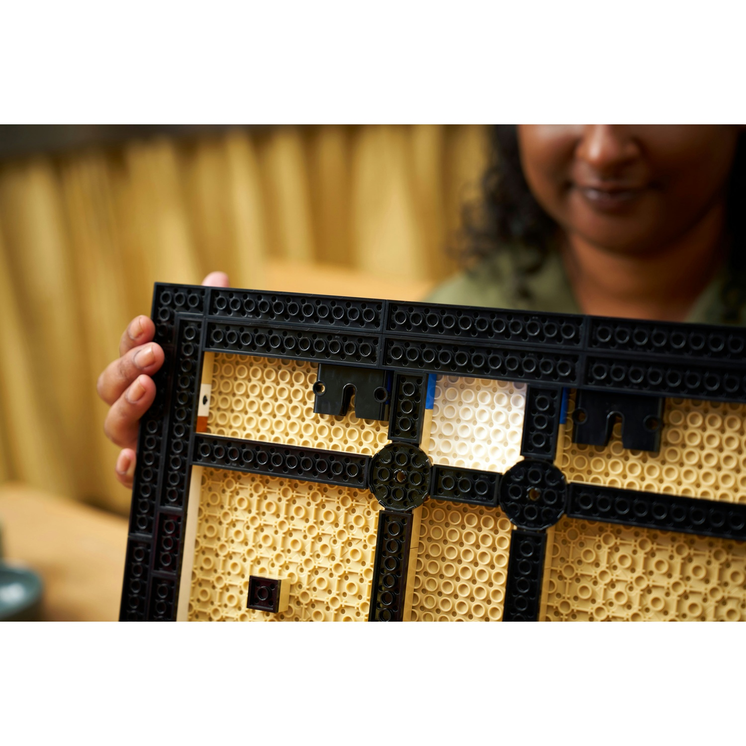 Una persona tiene in mano una cornice Lego nera con interno beige. La persona sorride. Lo sfondo mostra una tenda.