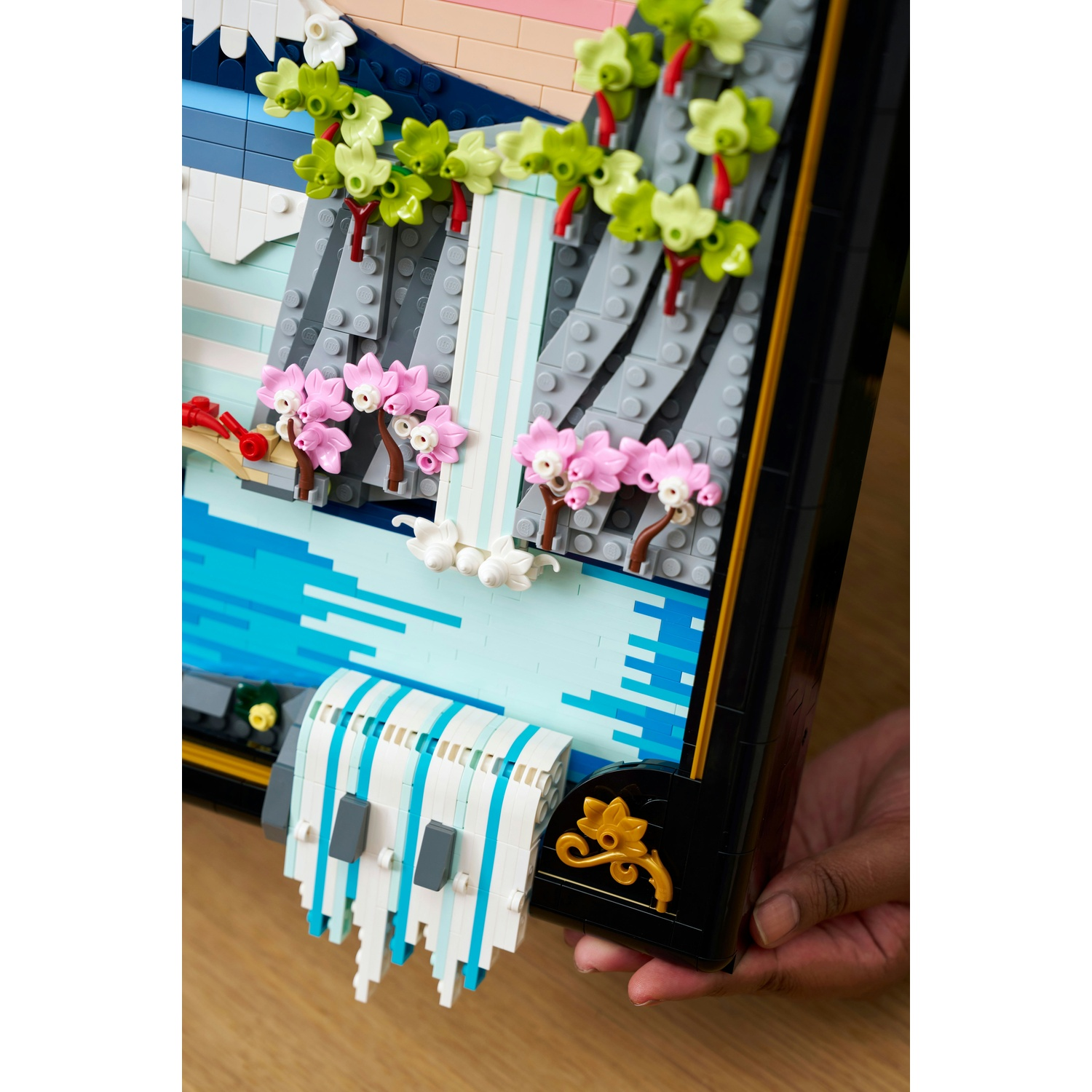 Scena Lego di una cascata con fiori rosa e fogliame verde. La scena è incorniciata in nero e tenuta da una mano su una superficie di legno.
