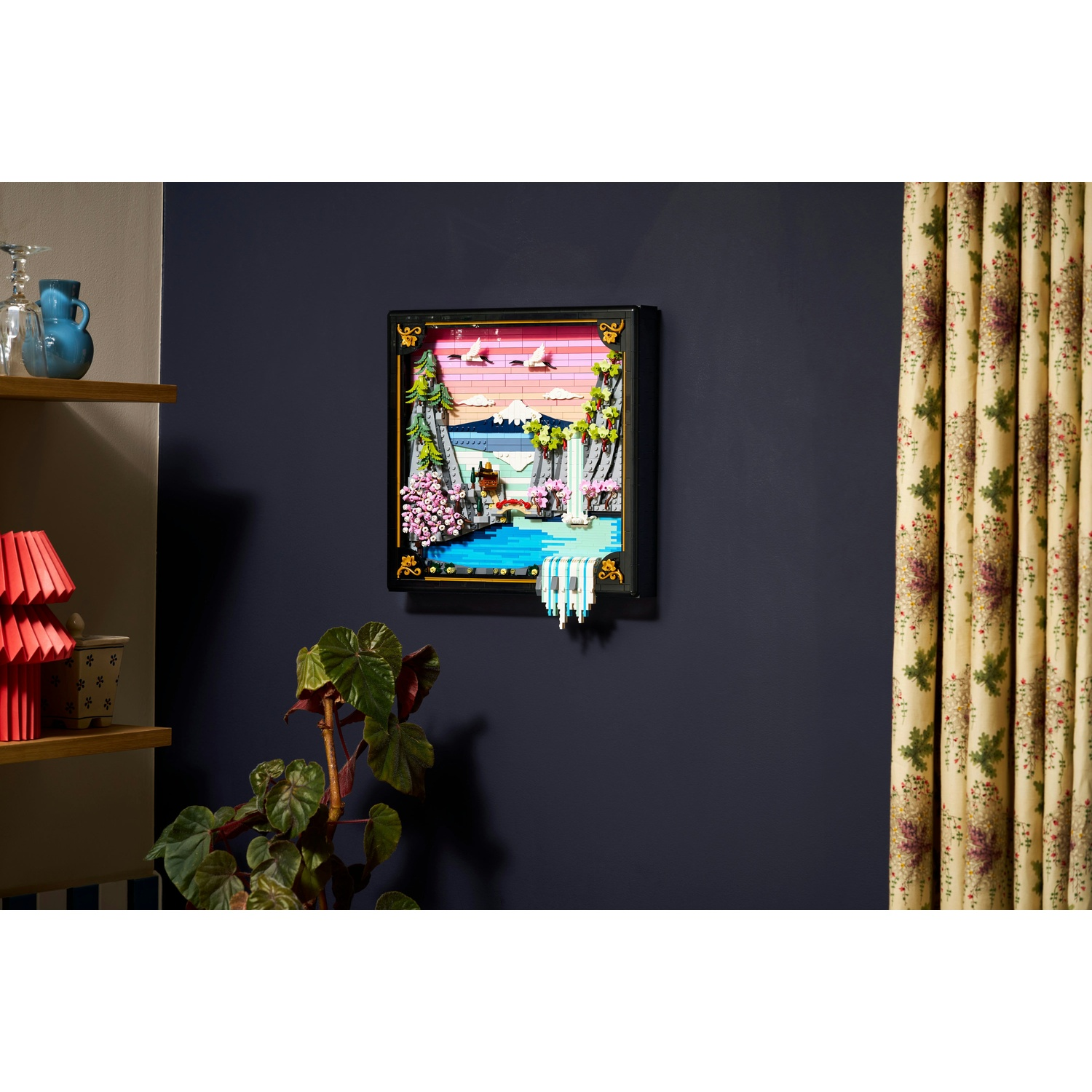 Un diorama Lego incorniciato raffigurante una scena di cascata con fiori rosa, uccelli e un cielo colorato, montato su una parete blu scuro.