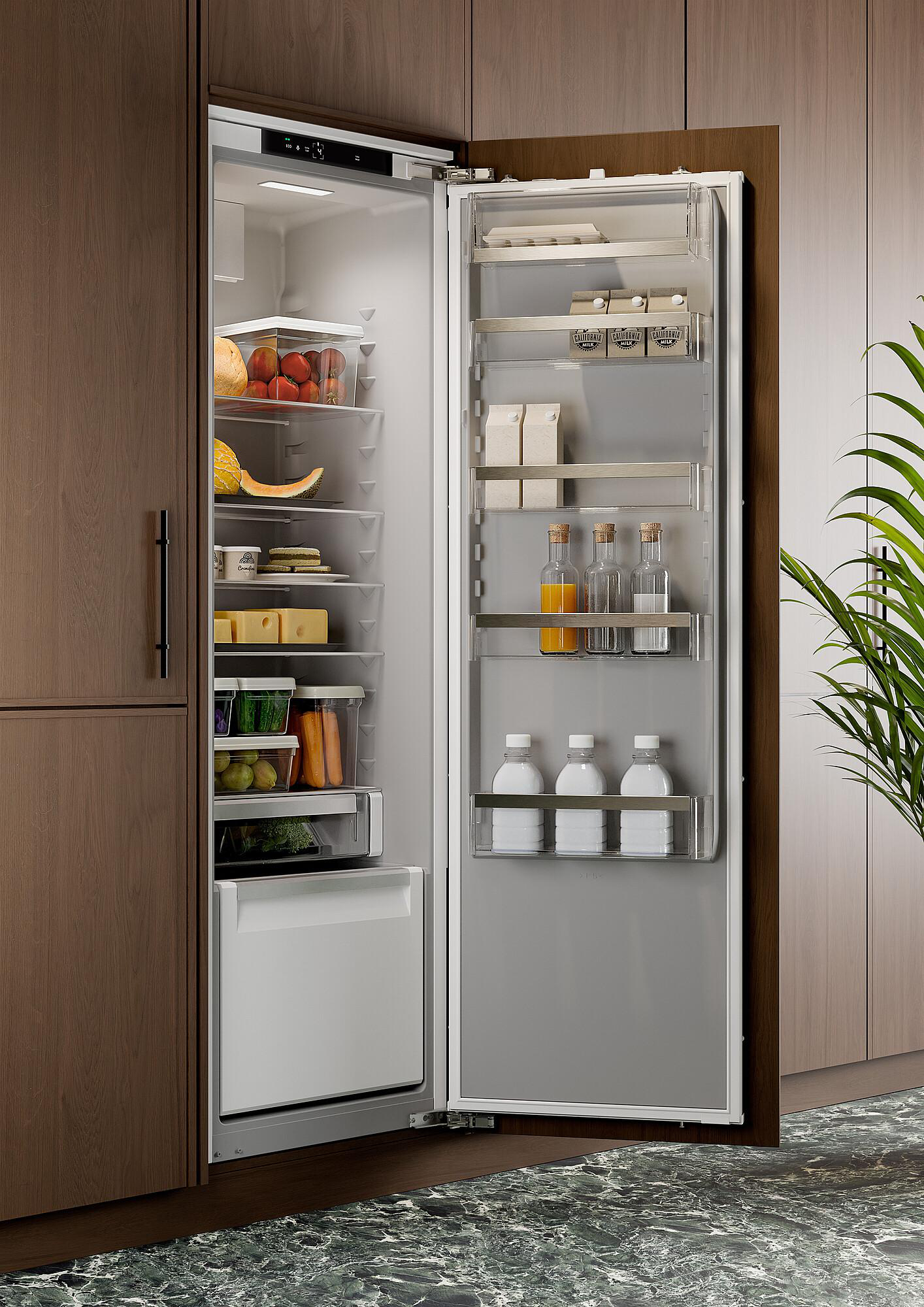 Offener Kühlschrank gefüllt mit Obst, Gemüse und Getränken. Er steht in einem Holzschrank. Rechts ist eine Pflanze.
