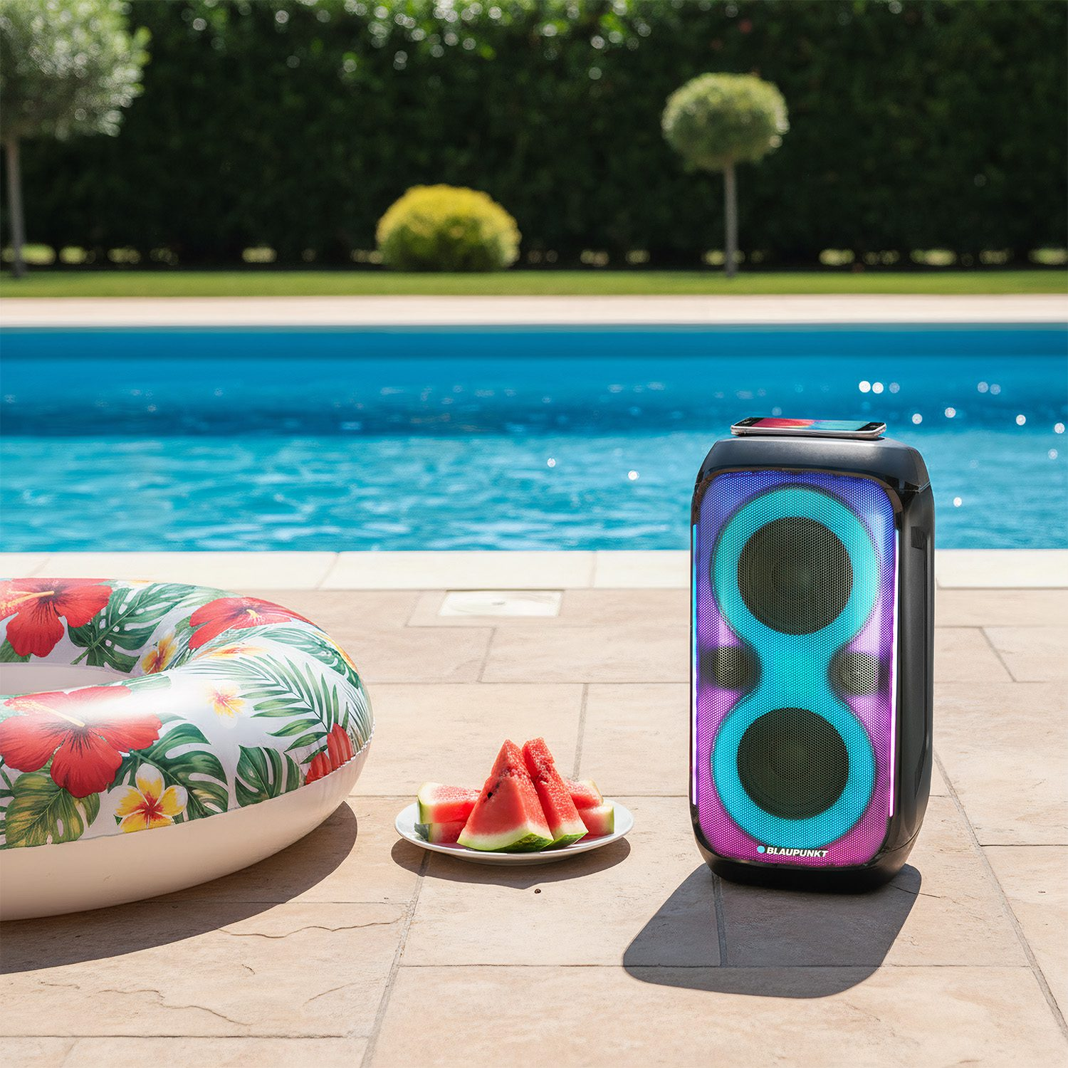 Głośnik, kawałki arbuza i nadmuchiwany koło obok basenu. Tło z drzewami i wodą w basenie.