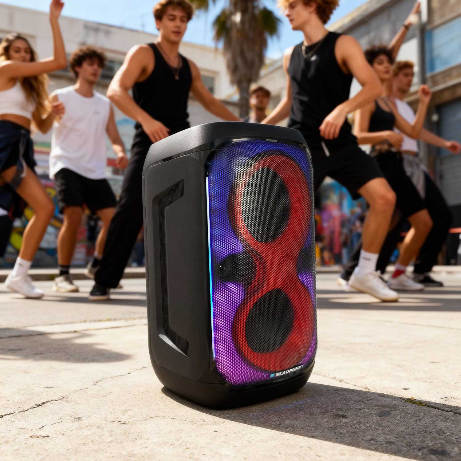 Czarny głośnik stoi na chodniku. Ludzie tańczą w tle. Głośnik ma czerwone, niebieskie i fioletowe światła.