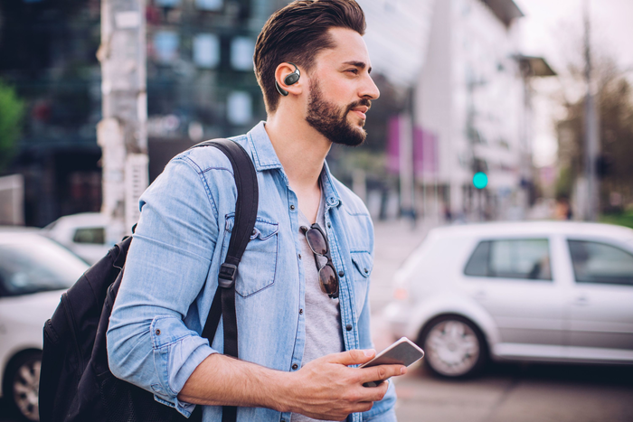 Ein Mann mit Bart, der Ohrhörer, ein Jeanshemd und einen Rucksack trägt und ein Telefon hält, steht auf einer Stadtstraße.