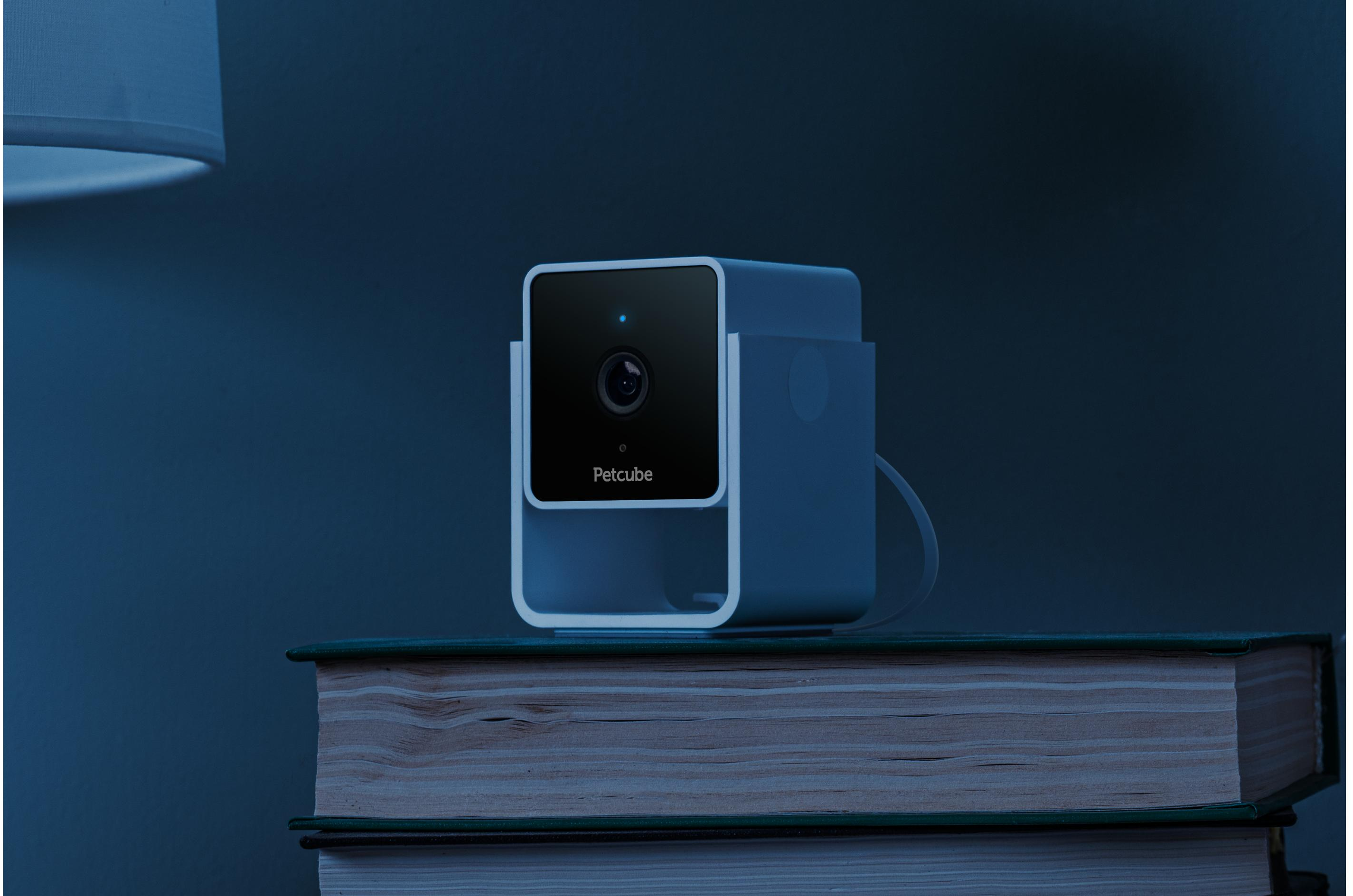 Una telecamera Petcube è sui libri. La telecamera è bianca e nera, con un obiettivo e una luce blu. Lo sfondo è blu.