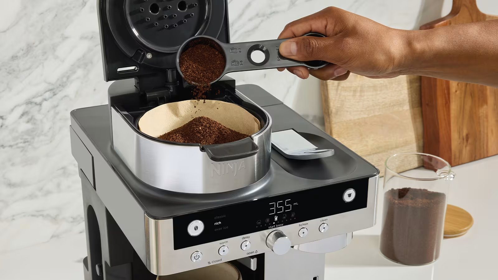 Una mano versa caffè macinato in un filtro di una macchina da caffè Ninja. La macchina è su un bancone con un tagliere di legno.