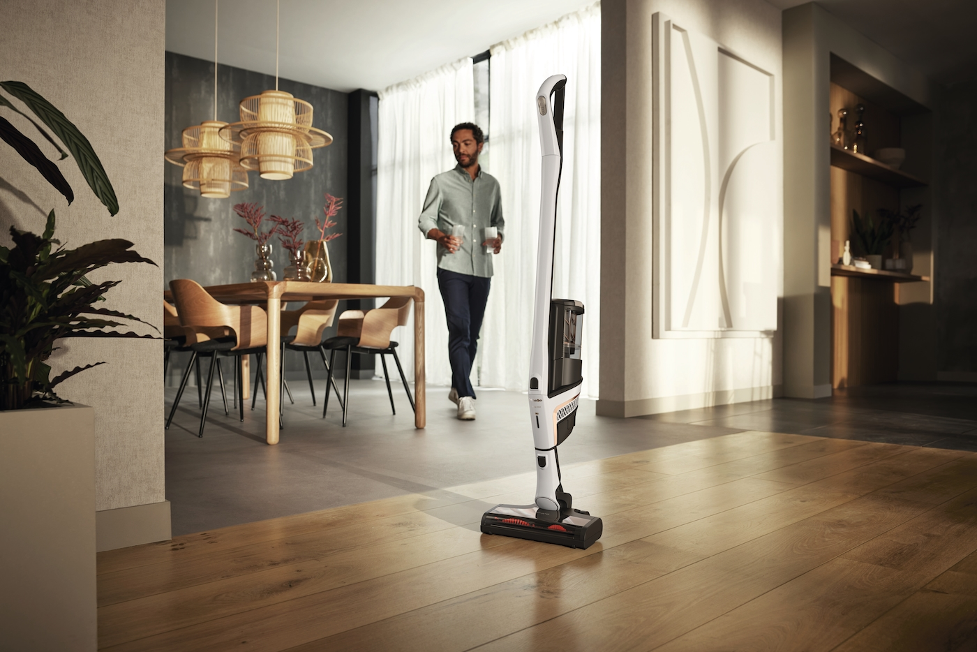 Ein Mann geht an einem weißen Staubsauger vorbei. Im Hintergrund sind ein Tisch, Stühle und Lampen zu sehen. Holzböden und Wände sind sichtbar.