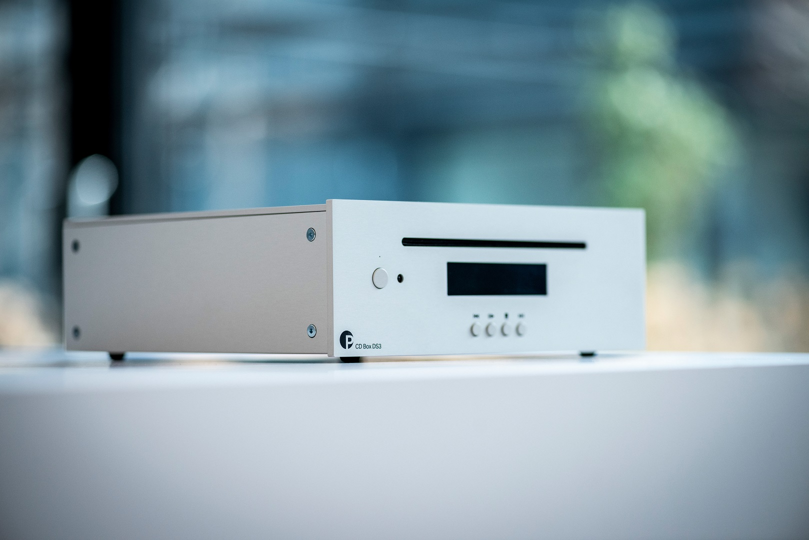 Ein silberner und weißer CD-Player steht auf einer weißen Oberfläche. Er hat ein Display, Knöpfe und das Markenlogo. Im Hintergrund ist ein verschwommenes Fenster.