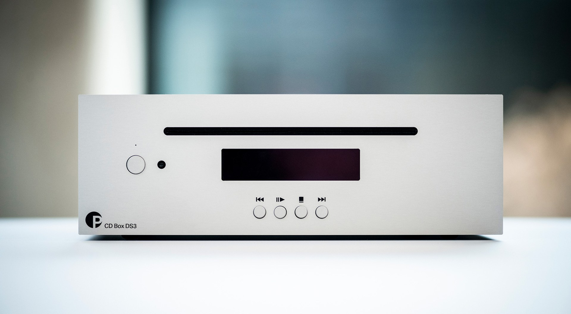 Silberner CD-Player auf weißer Oberfläche. Hat ein Display, Knöpfe und einen Schlitz. Der Hintergrund ist unscharf.