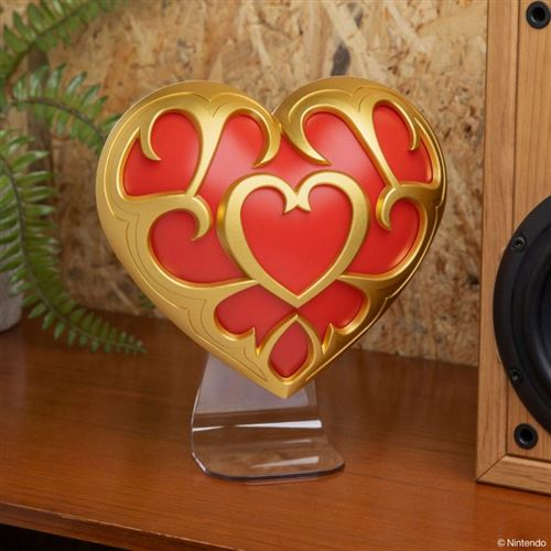 Un objeto decorativo en forma de corazón con adornos dorados y centro rojo se exhibe sobre una superficie de madera, junto a un altavoz.