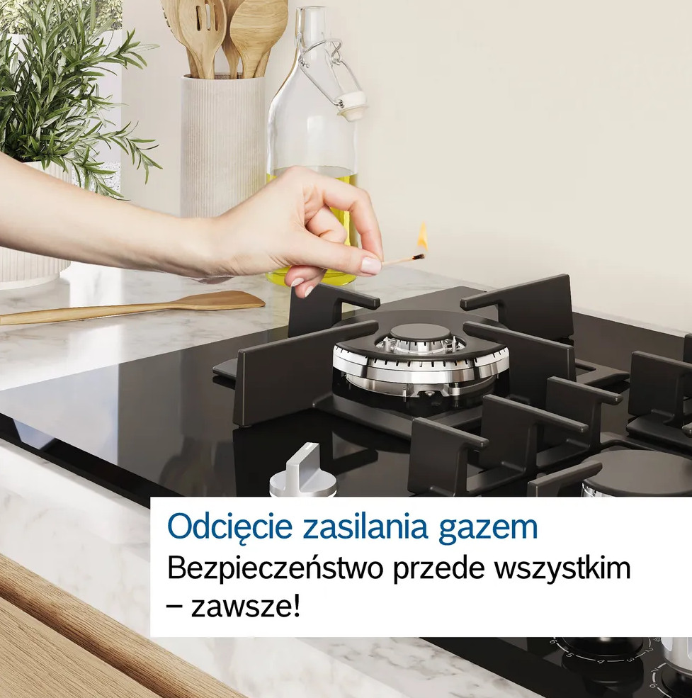 Ręka zapala zapałkę nad czarną kuchenką gazową. Tło obejmuje przybory kuchenne, szklaną butelkę i biały blat.