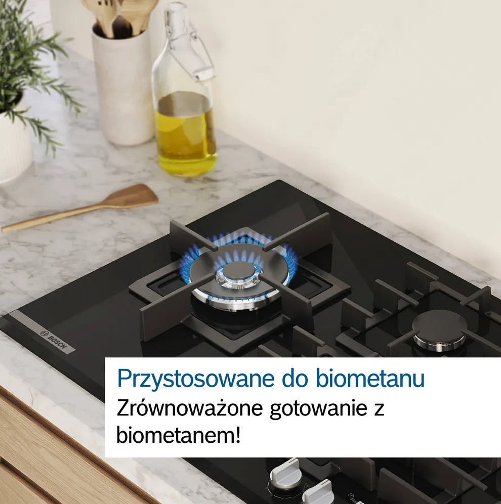 Czarna płyta gazowa z niebieskim płomieniem jest włączona. Za nią stoi butelka oleju i przybory kuchenne.