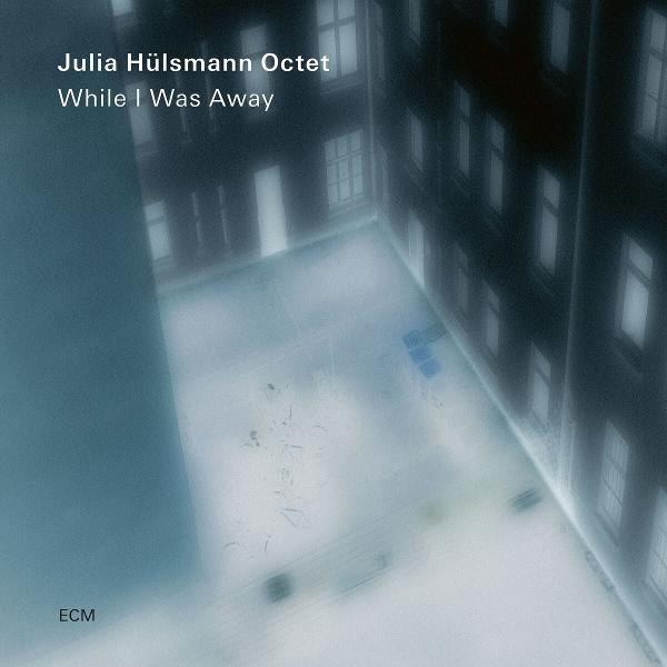 Albumcover mit dem Titel 'While I Was Away'. Gebäude mit Fenstern rechts. Blau- und Weißtöne, ein leerer Raum in der Mitte.